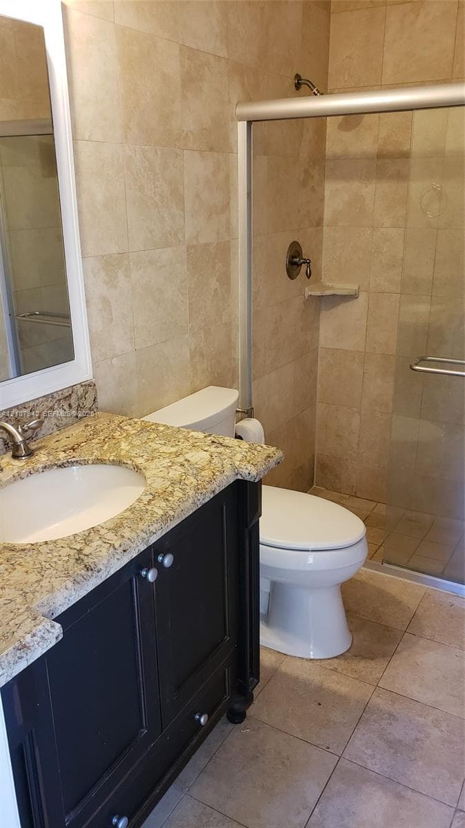 Bathroom featuring a dark-finish vanity with a stone-surfaced countertop, an undermount sink, a walk-in shower with a sliding glass door, and neutral-toned wall and floor tile