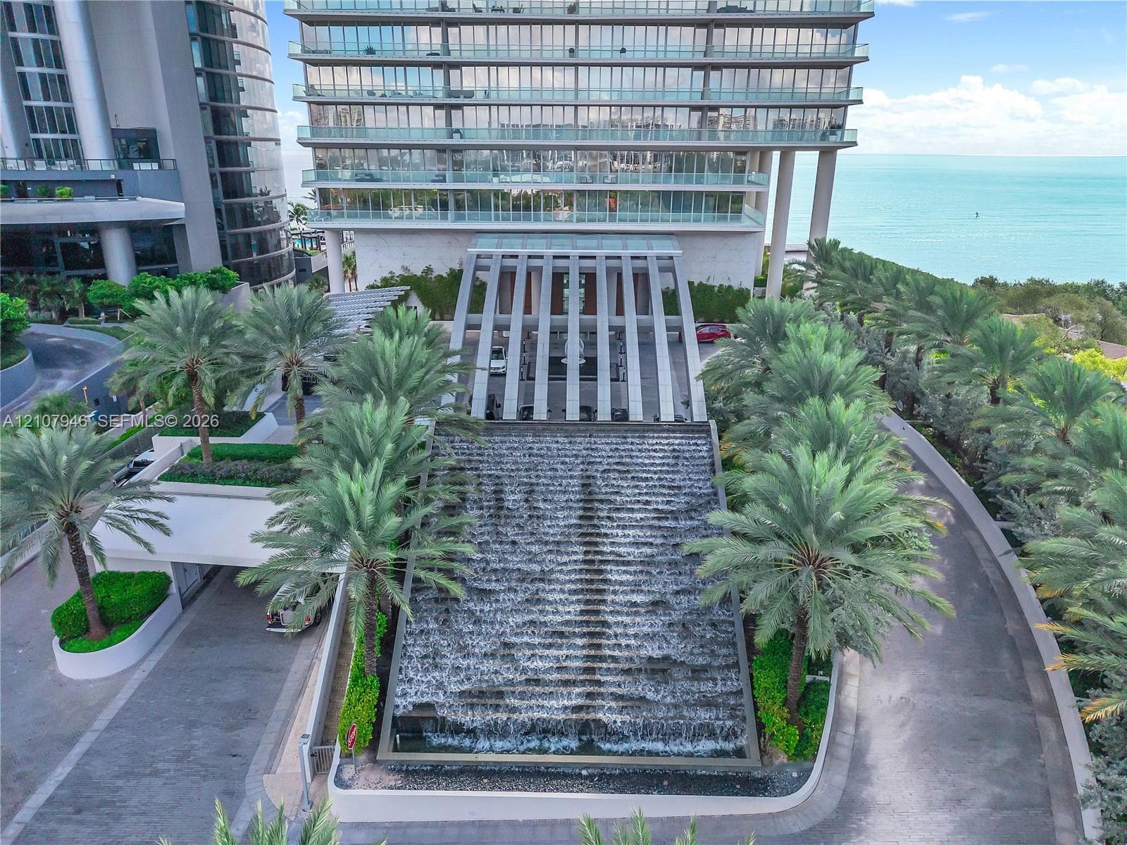 High-rise residential building featuring a tiered waterfall feature, a covered porte-cochère with structural pillars, landscaped palm trees, and glass-railed balconies