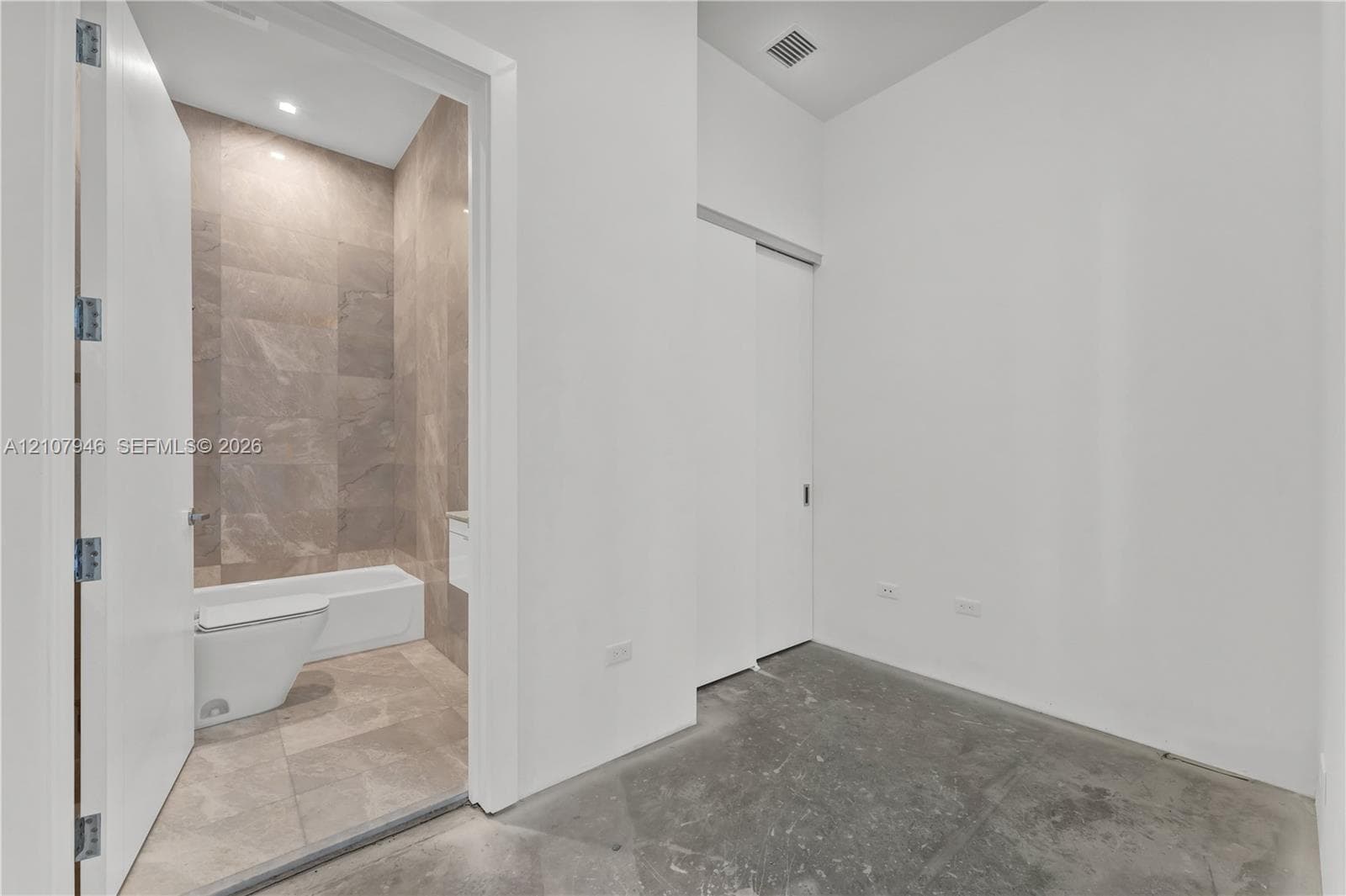 Interior space featuring polished concrete flooring and a sliding door, revealing a bathroom finished with large-format tile flooring and wall surfacing, a white bathtub, and a wall-mounted toilet