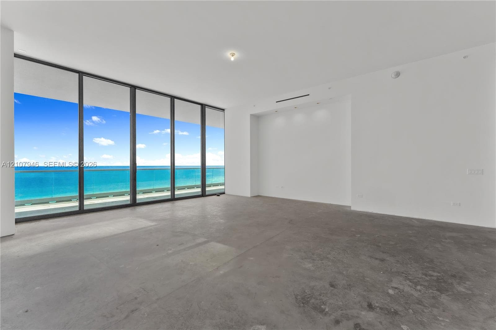 Floor-to-ceiling windows, full-height glass sliding doors, private glass-railed balcony, polished concrete flooring, and ocean backdrop