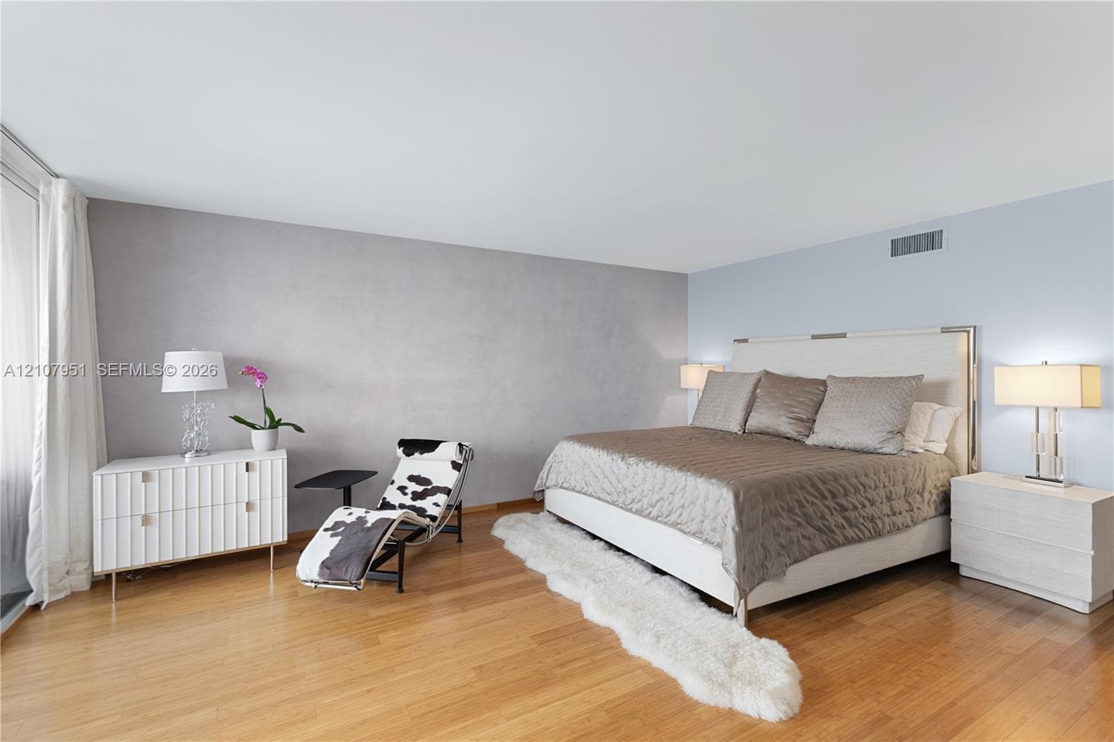 Spacious primary bedroom showcasing warm wood-finish flooring, a generous bed layout with upholstered headboard, dual nightstands with integrated lighting, and abundant natural light from large windows