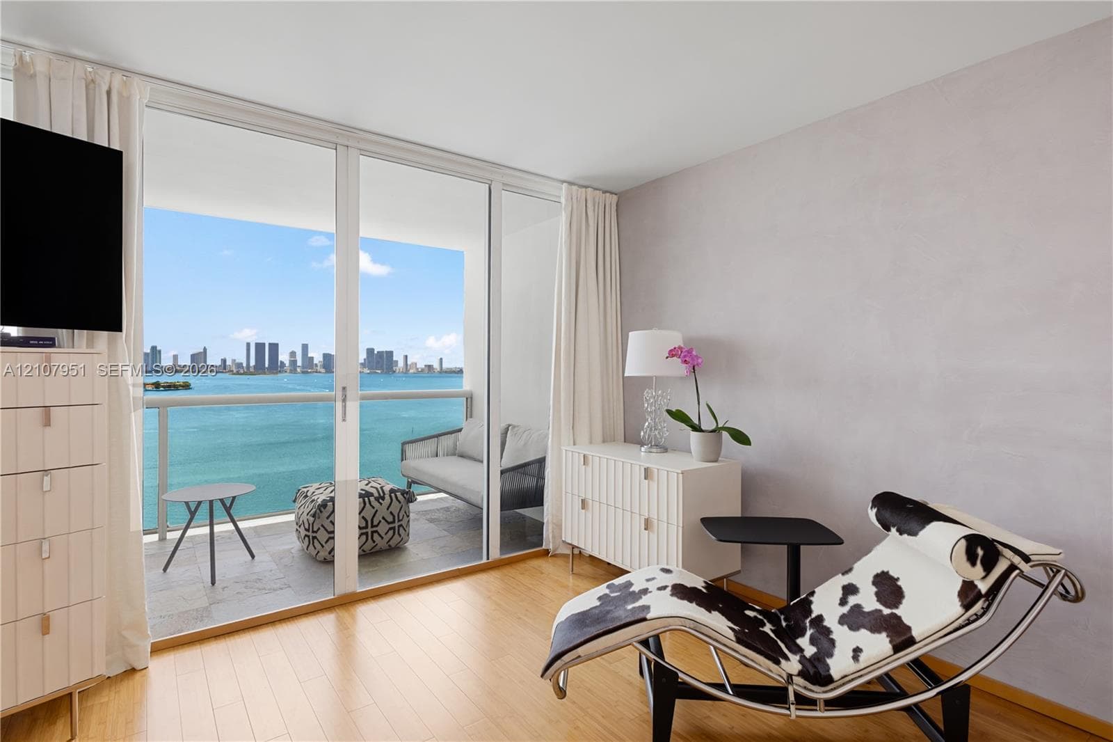 Floor-to-ceiling glass sliding doors opening to a balcony with metallic railing, wood-finish flooring, and expansive water and skyline backdrop