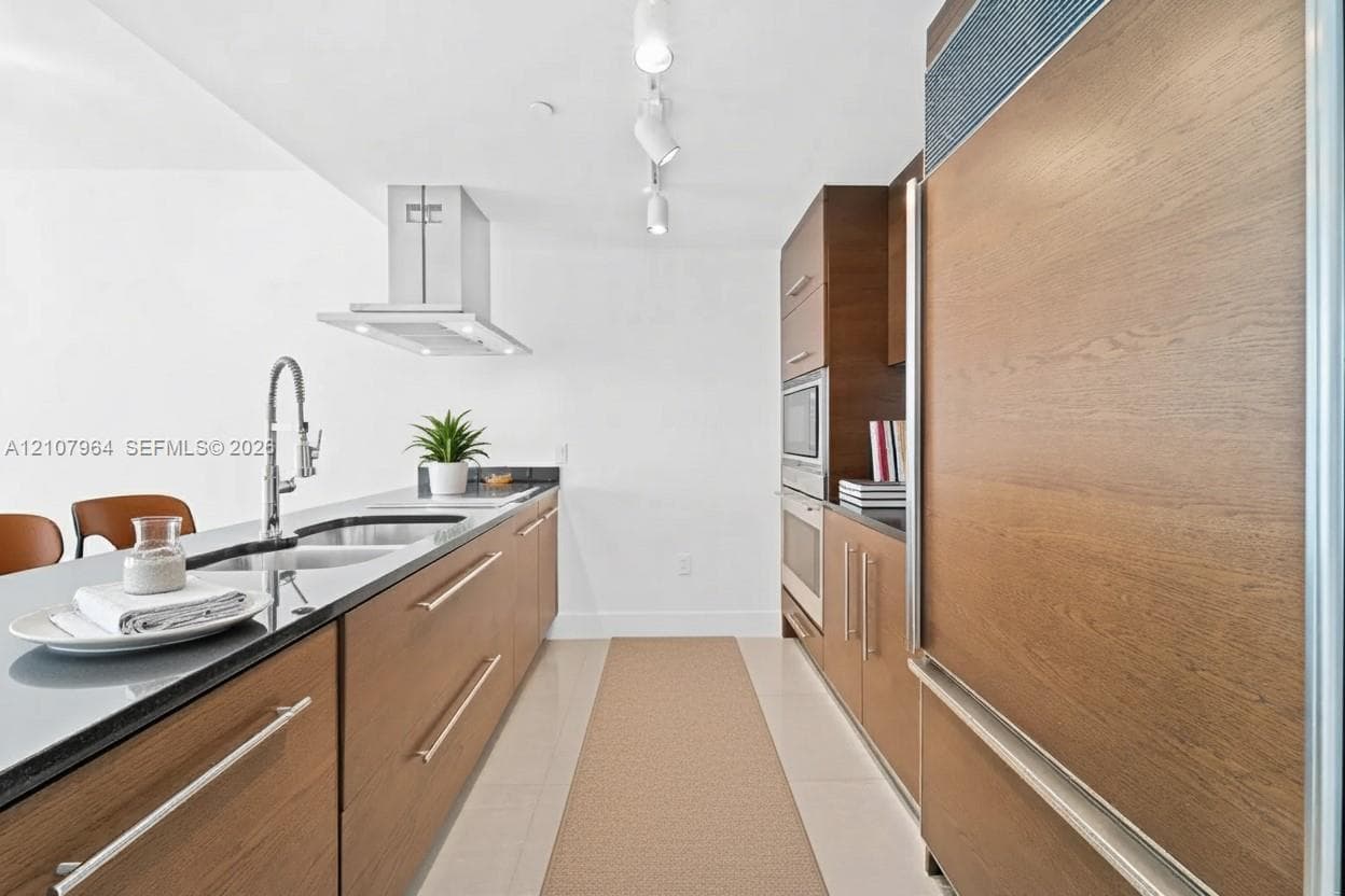 Kitchen featuring wood-finish cabinetry, stainless steel panel-ready refrigerator, built-in wall oven and microwave, dark stone countertops with an integrated sink, and stainless steel range hood