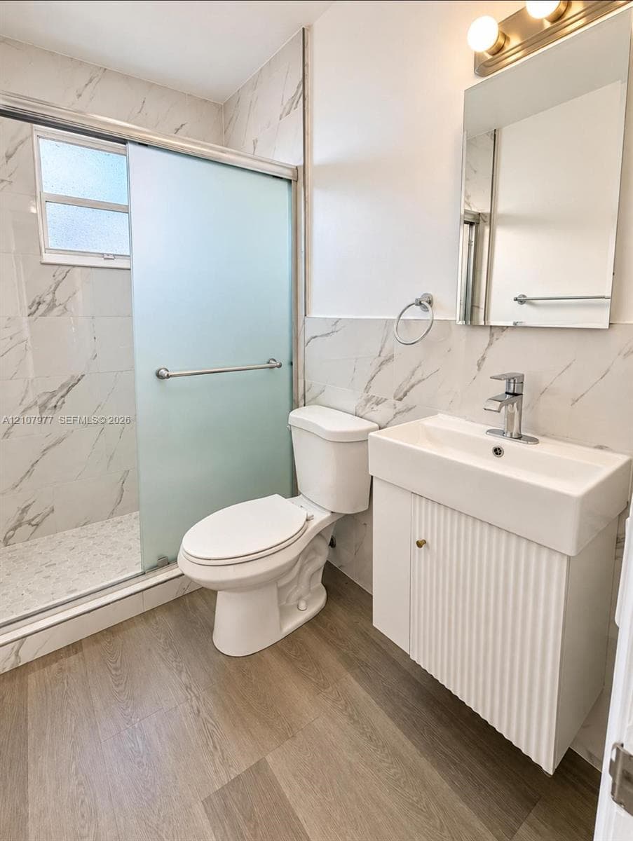 Bathroom featuring stone-finish wall tile, fluted-front vanity with a rectangular sink, walk-in shower with frosted sliding glass doors, and wood-finish flooring