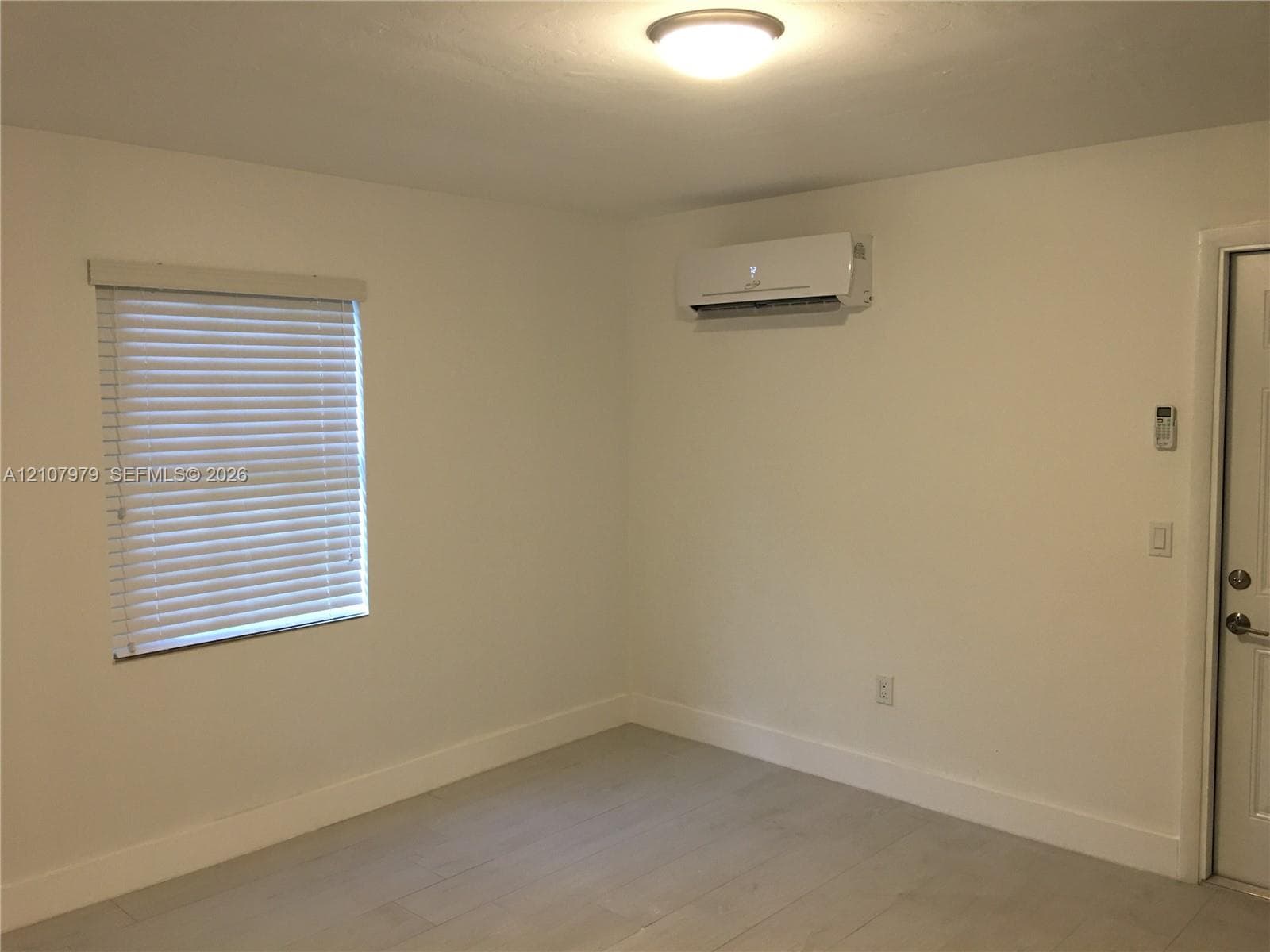 Interior space featuring wood-finish flooring, a wall-mounted ductless mini-split air conditioning unit, a window with horizontal blinds, and a flush-mount ceiling light fixture