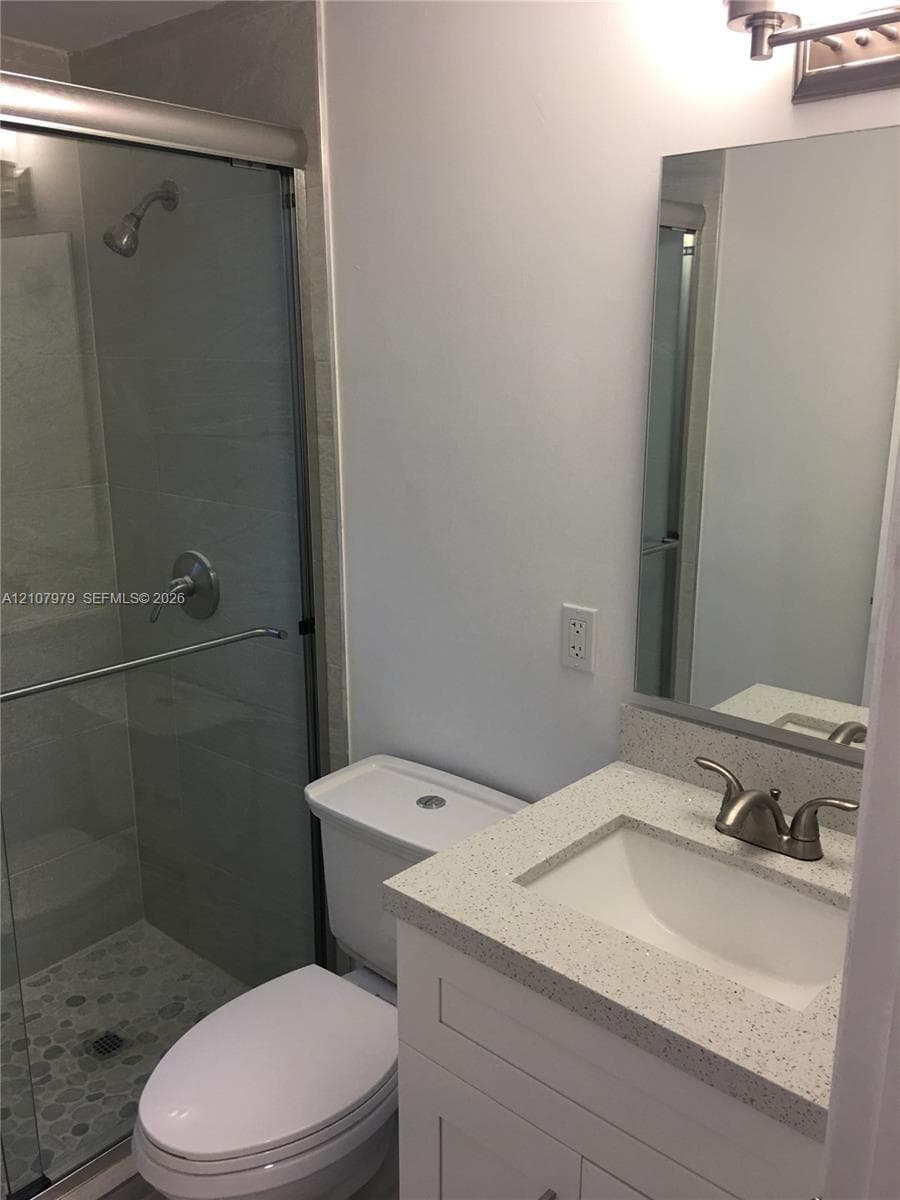 Bathroom featuring a stone-composite countertop, white vanity cabinet, undermount sink with dual-handle faucet, and walk-in shower with pebble-tile flooring and glass enclosure