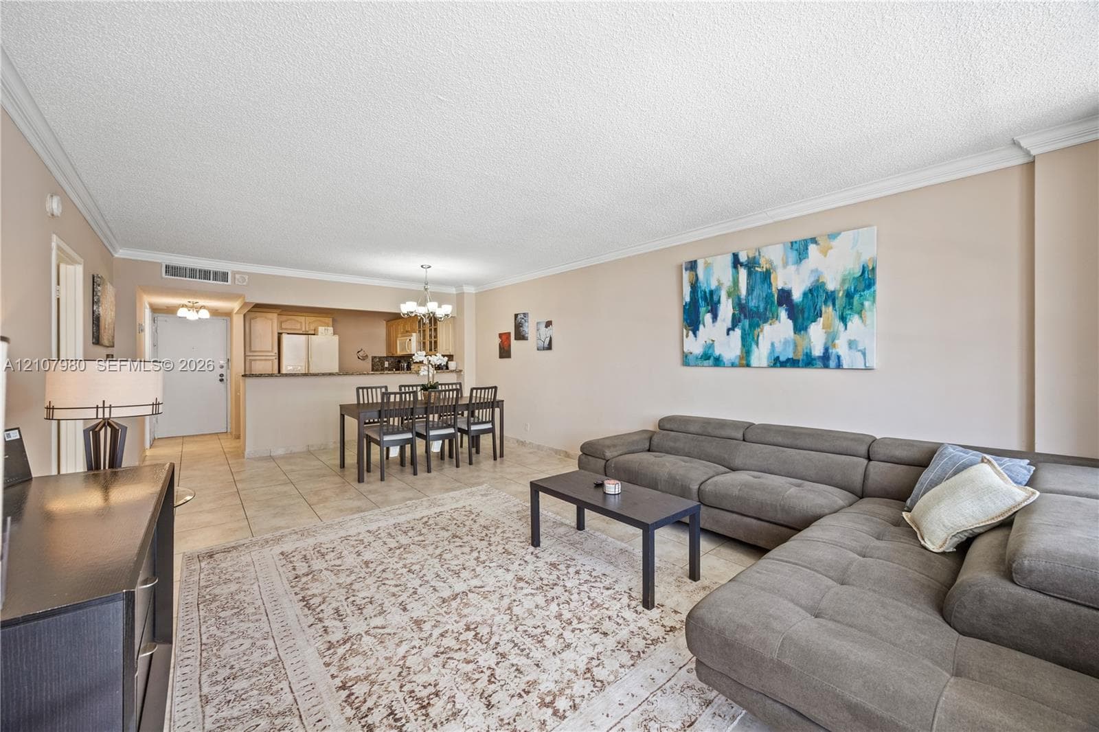 Open-concept interior featuring large-format neutral floor tiles, crown molding, a kitchen pass-through, and a decorative chandelier