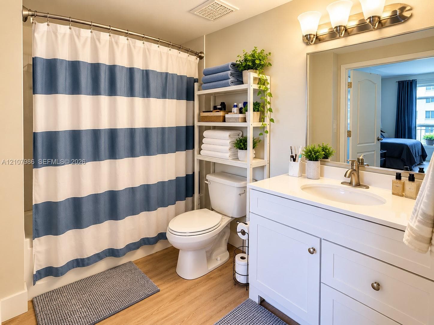 Bathroom featuring wood-finish flooring, a white vanity with a solid-surface countertop and integrated sink, a brushed nickel faucet, a large frameless mirror, and a tri-light fixture
