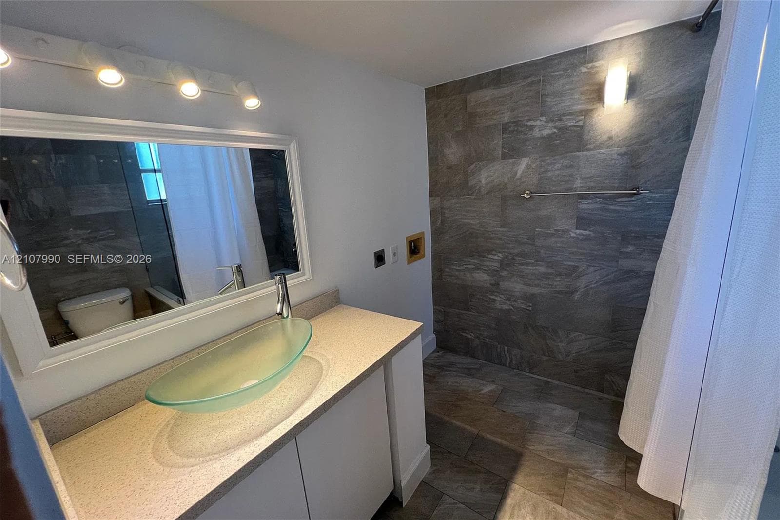 Bathroom vanity featuring a speckled countertop, glass vessel sink, and four-light wall-mounted fixture, with stone-tiled shower surround
