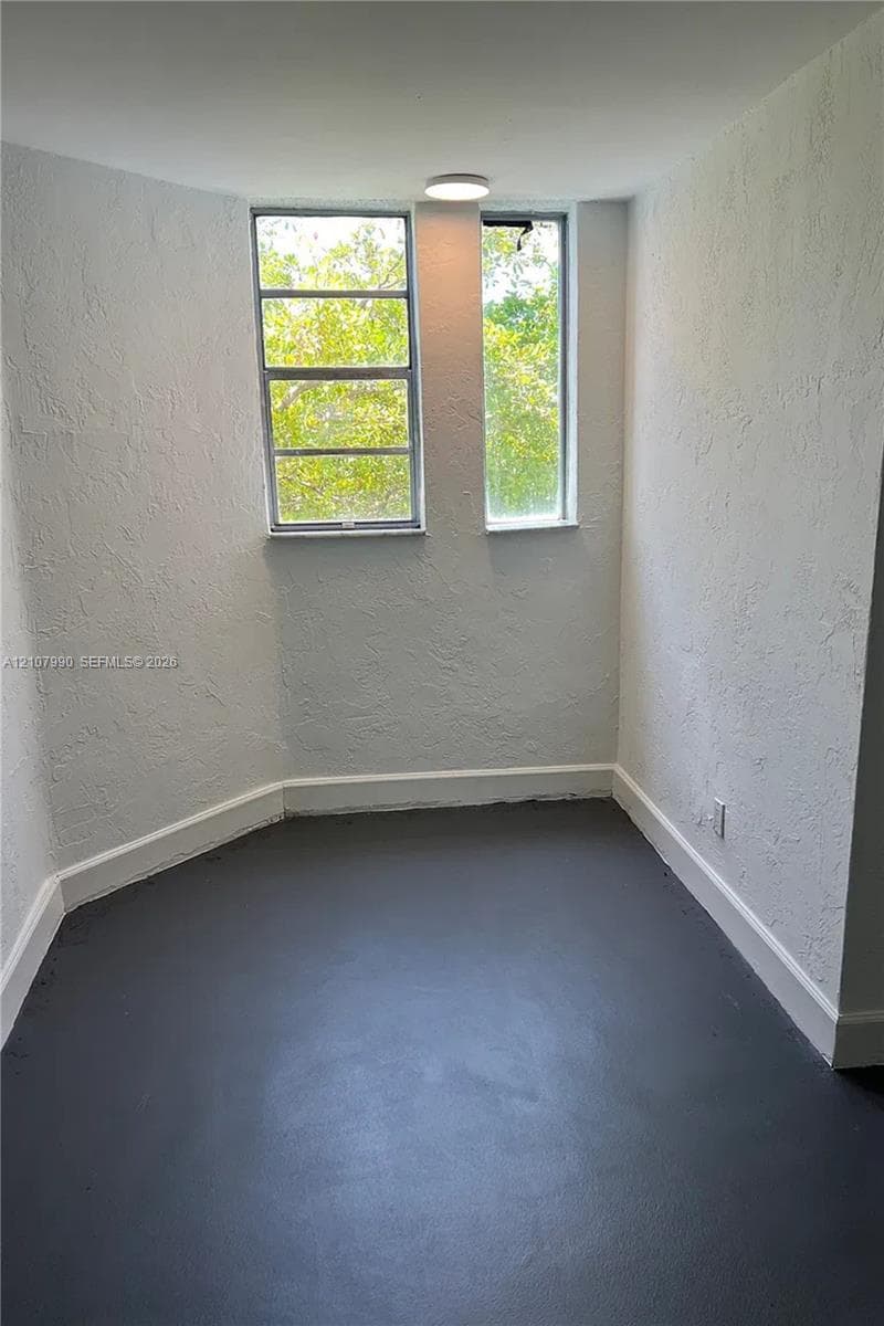 Multi-faceted room with textured walls, painted concrete flooring, two multi-pane windows, and a flush-mount ceiling light fixture