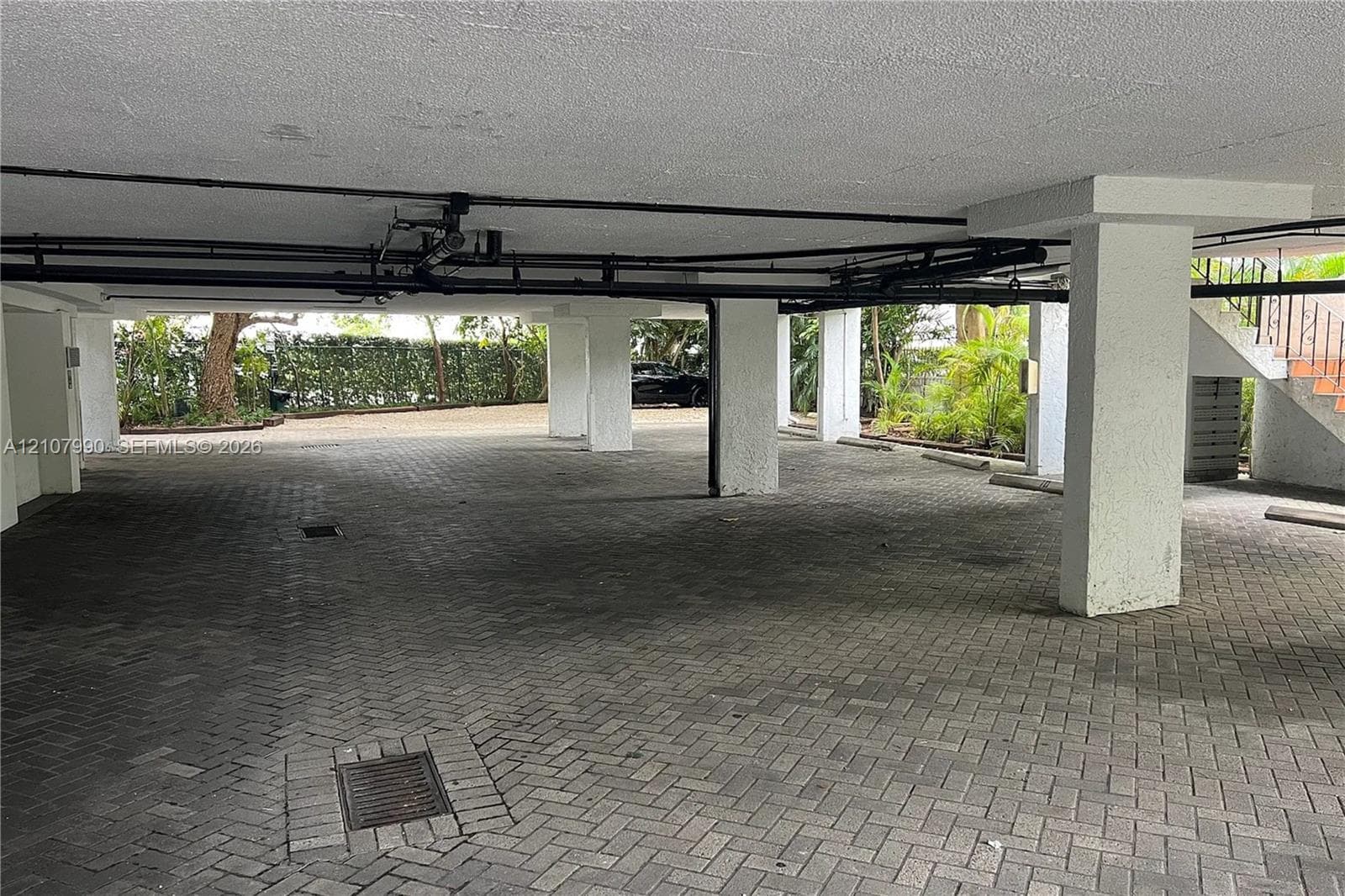 Covered parking area featuring herringbone-pattern paver flooring, structural concrete support pillars, and integrated drainage