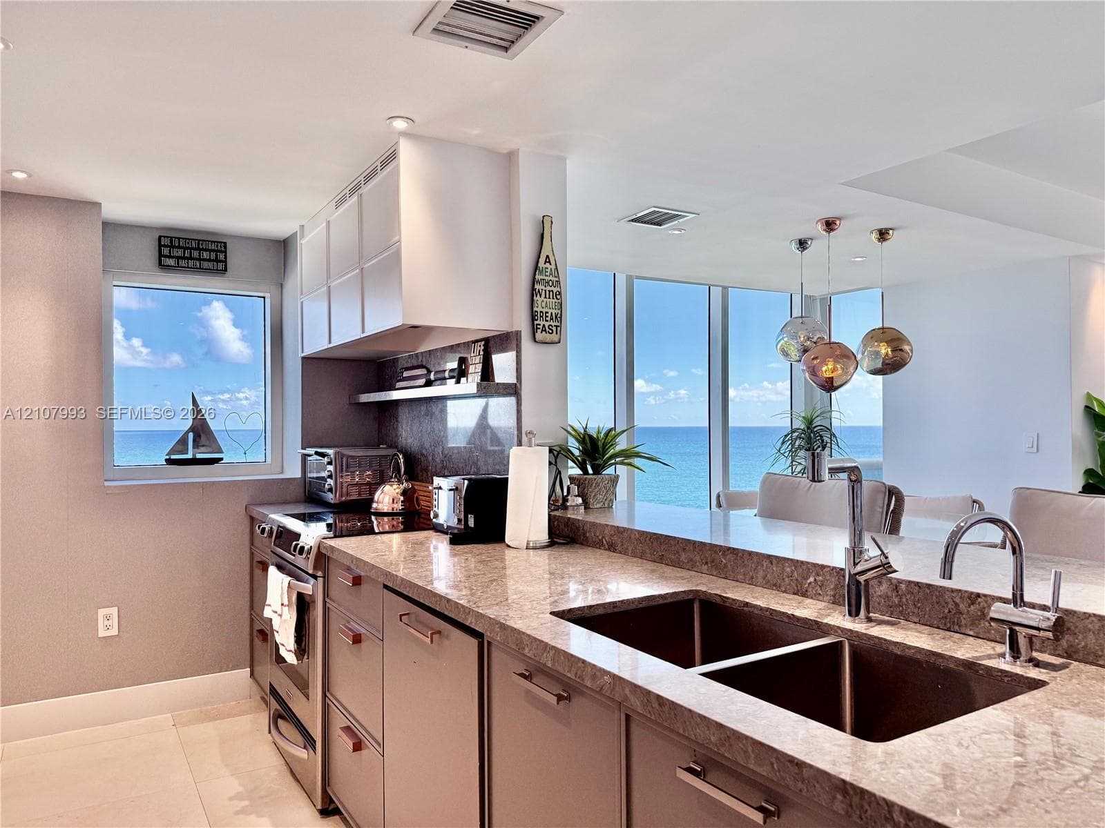 Kitchen featuring flat-panel cabinetry, stone countertops, stainless steel appliances, double-basin undermount sink with high-arc faucet, and floor-to-ceiling windows with coastal scenery
