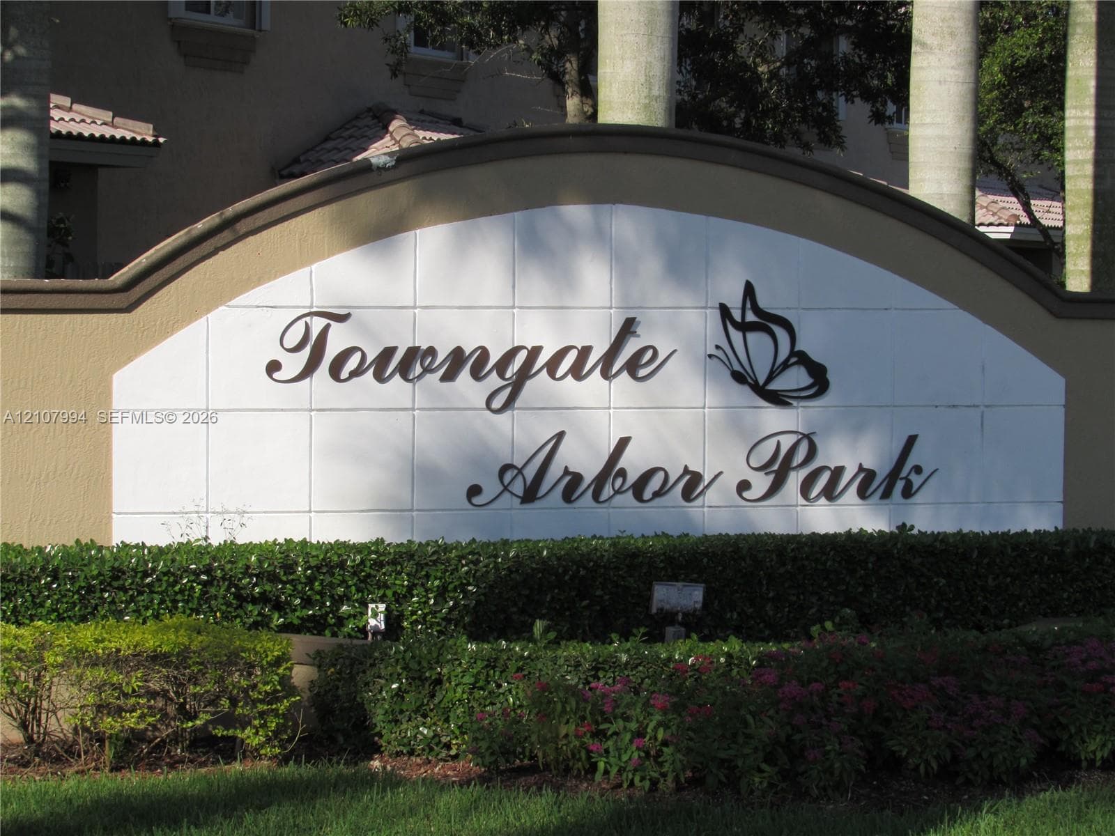 Arched monument signage featuring dimensional lettering and decorative metal butterfly detailing, set against a concrete-tiled backdrop with manicured shrubbery and palm tree landscaping