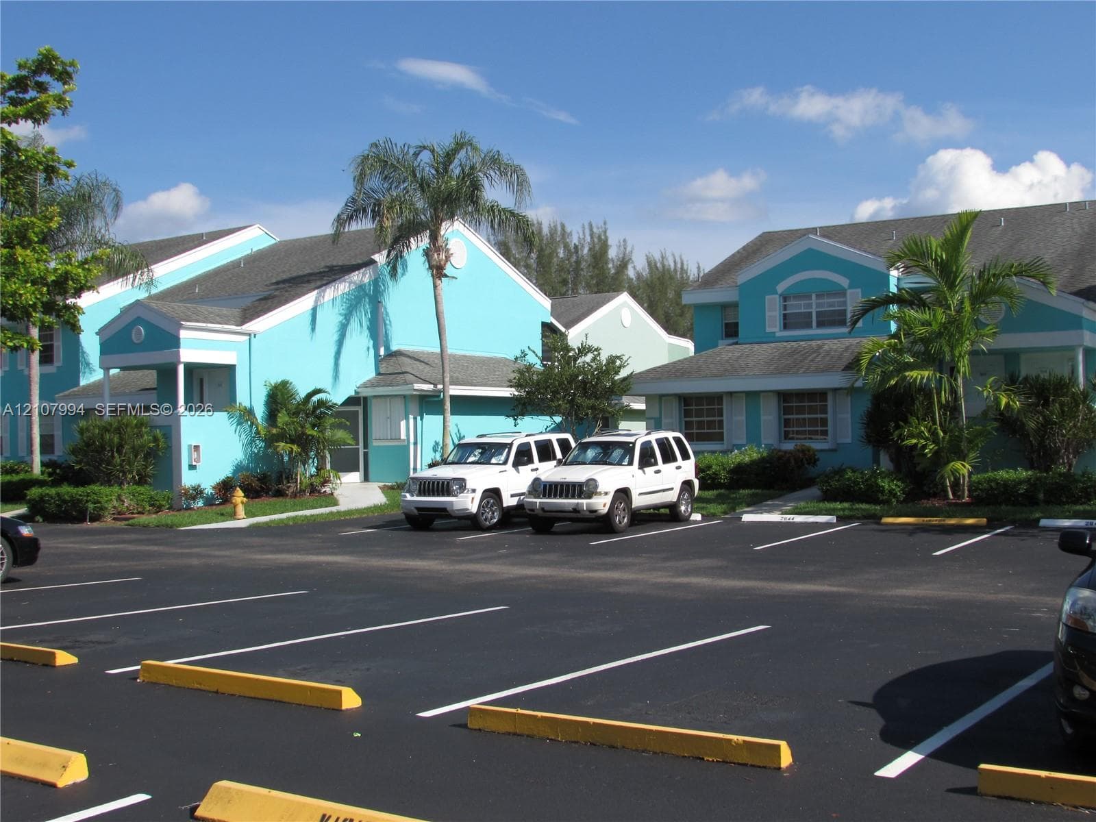 Multi-family residential complex featuring blue exterior siding, gabled rooflines, paved surface parking, and established palm landscaping