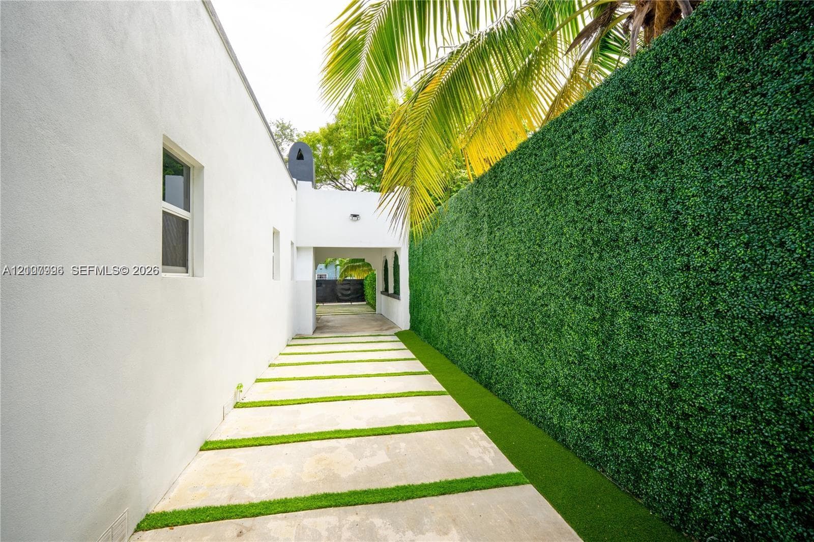 Exterior side walkway featuring concrete pavers with artificial turf strips and an artificial hedge wall