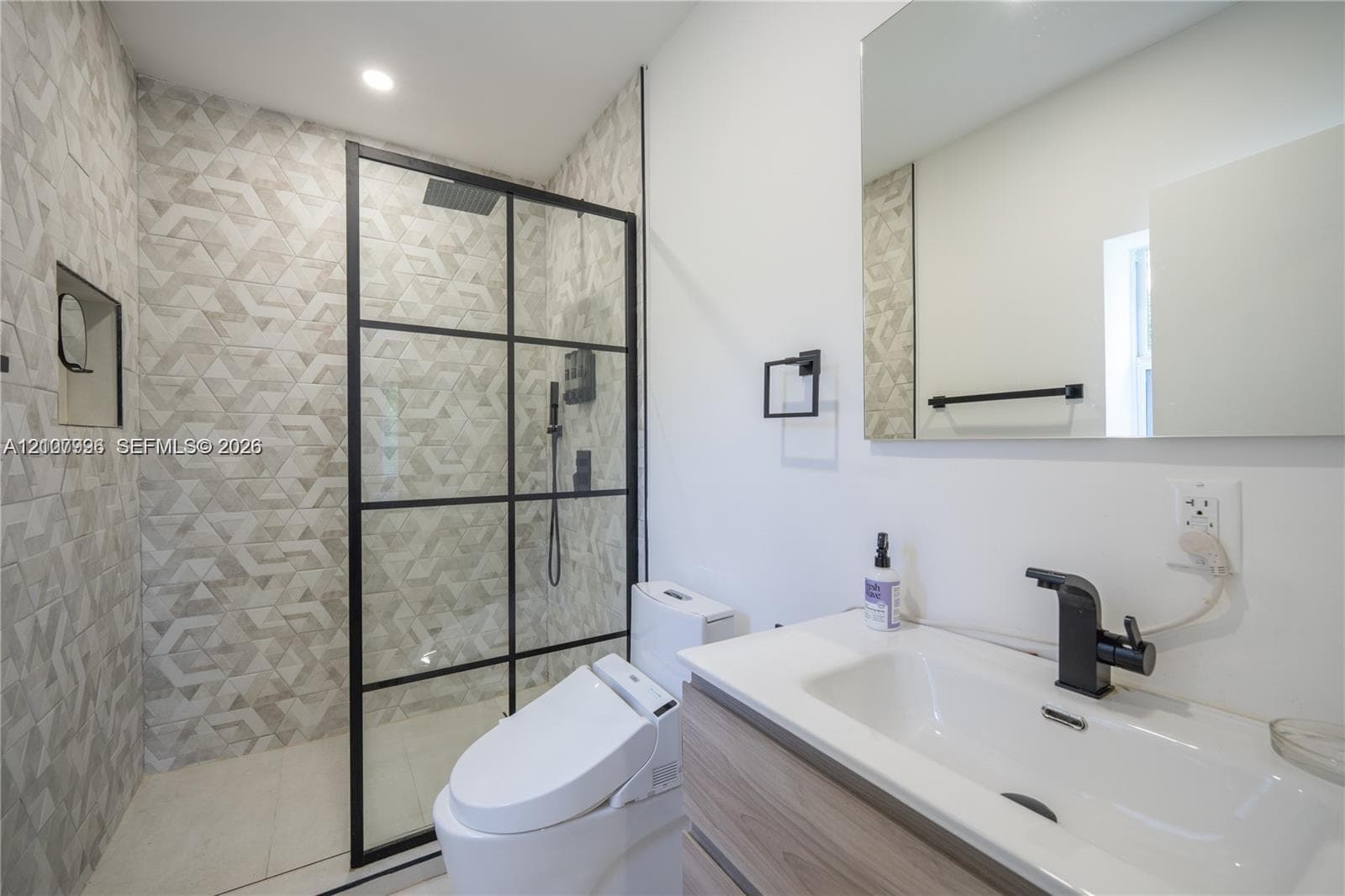 Walk-in shower featuring a grid-pattern black frame, geometric wall tiling, and a recessed niche, complemented by a wood-finish vanity with a matte black faucet and a wall-mounted mirror