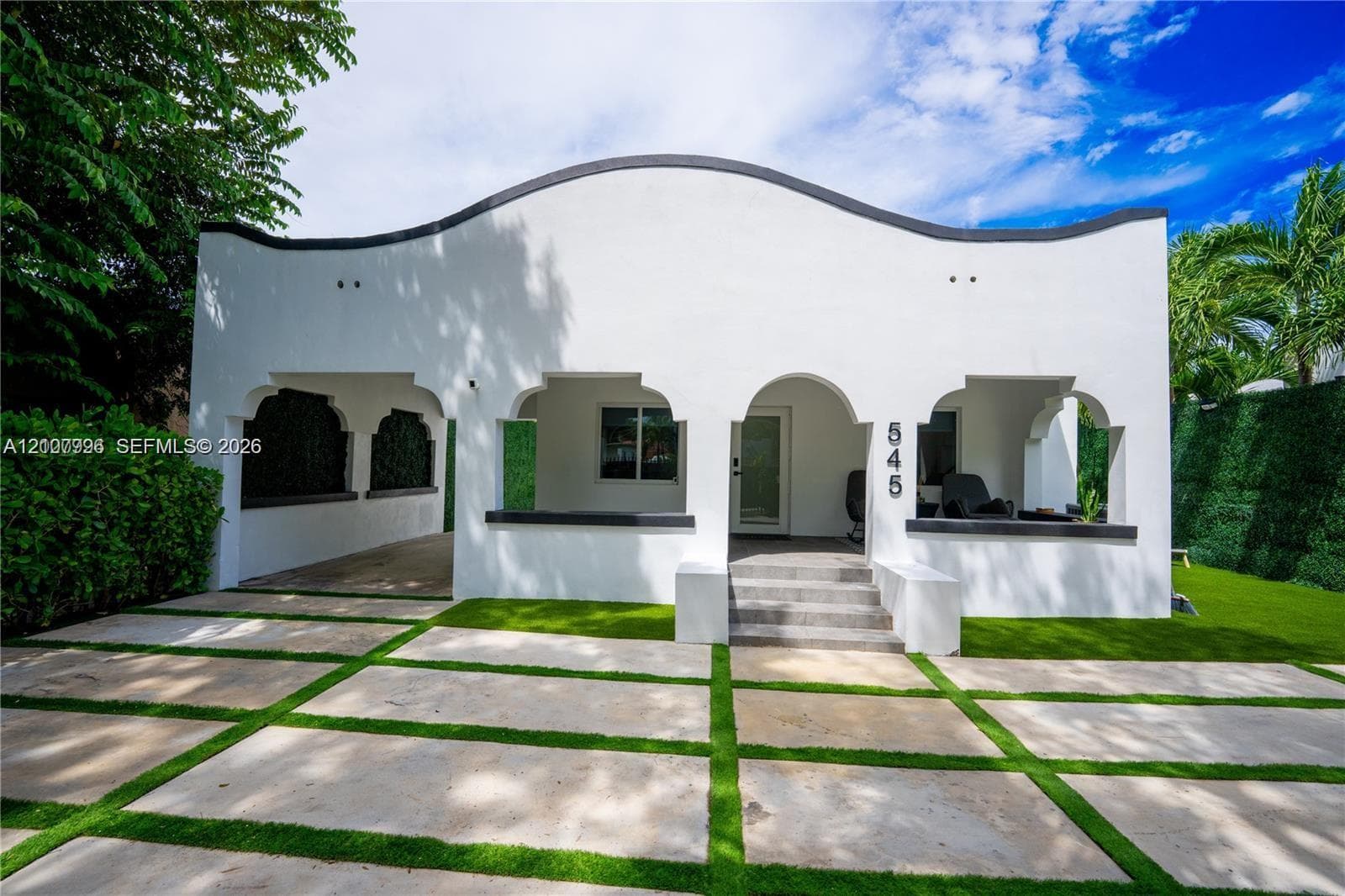 Spanish Colonial-style exterior featuring a scalloped roofline, arched portico, covered carport, and concrete paver driveway with integrated artificial turf