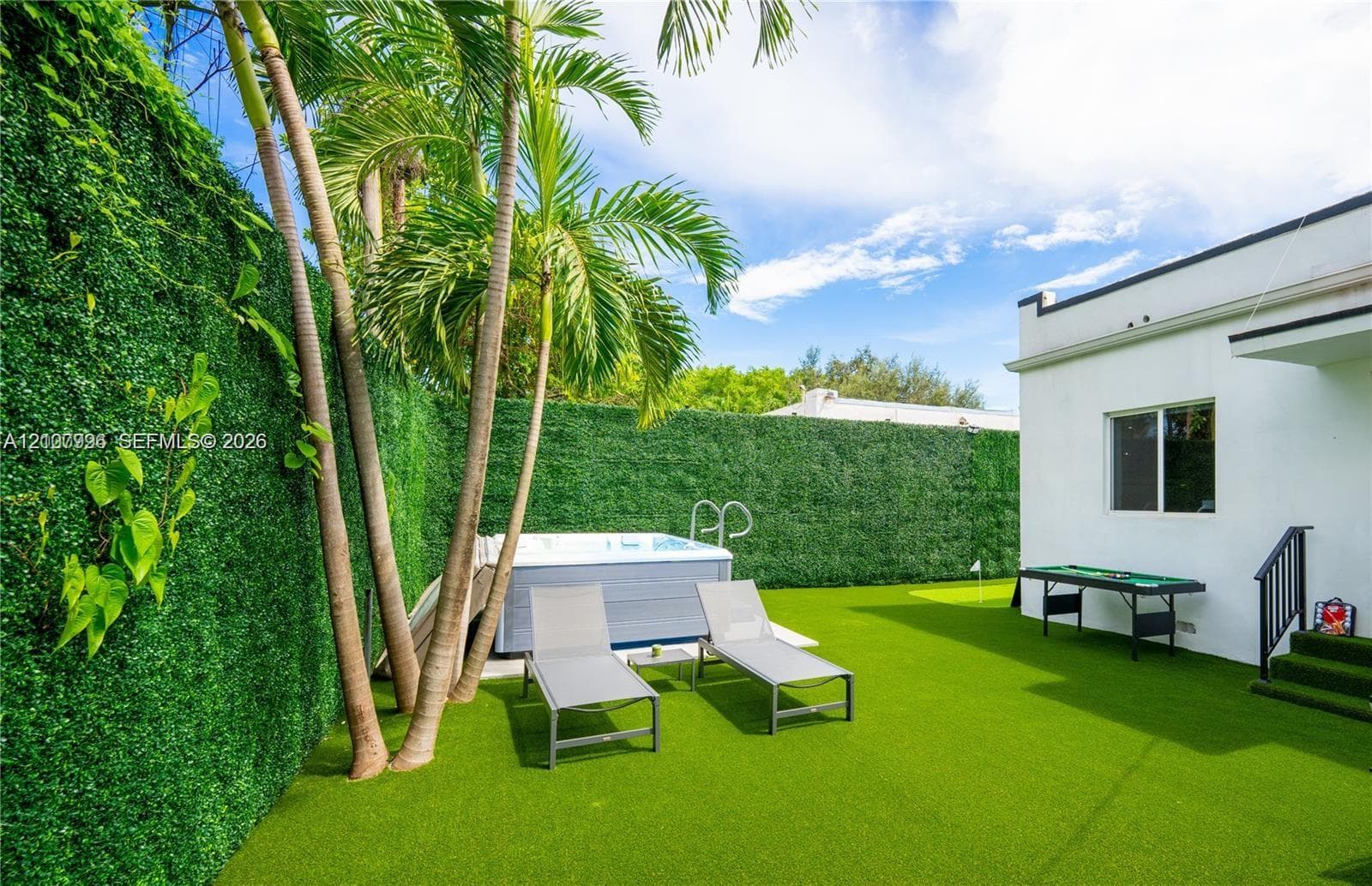 Backyard featuring synthetic turf, perimeter privacy hedging, a built-in spa, mature palm trees, and a putting green