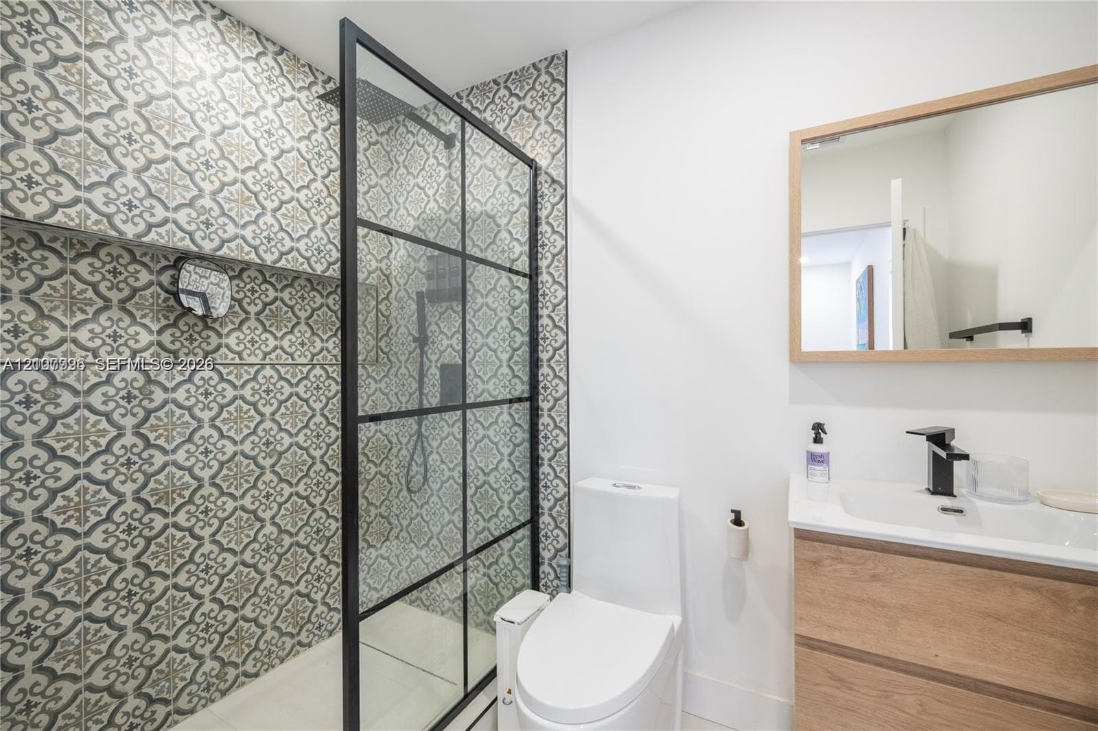 Walk-in shower with black-frame glass enclosure and patterned wall tile, wood-finish vanity with integrated sink, and wall-mounted rectangular mirror