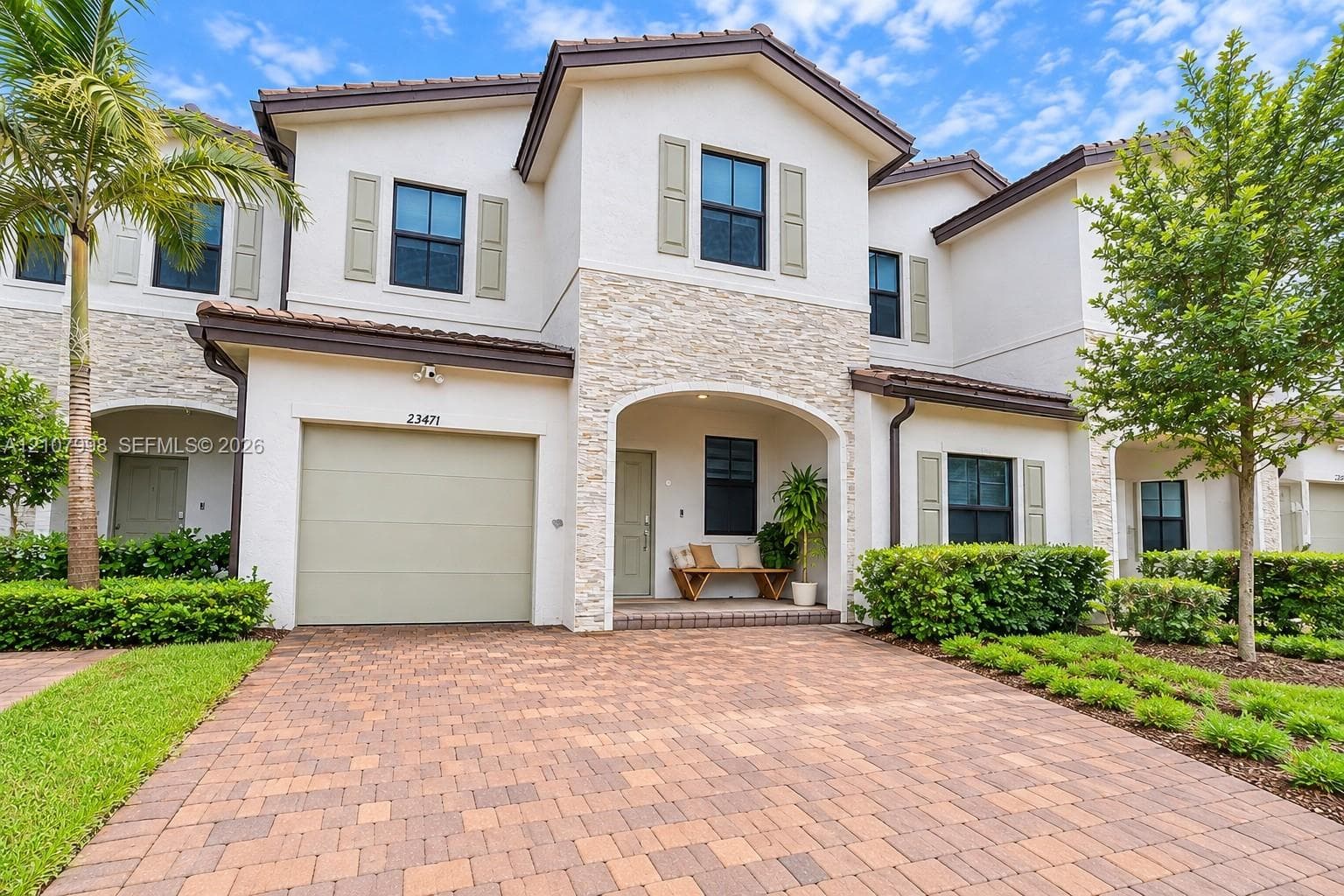 Two-story exterior featuring light-toned stucco, decorative stone veneer accents, a paver driveway, an arched covered entryway, and shutter-framed windows