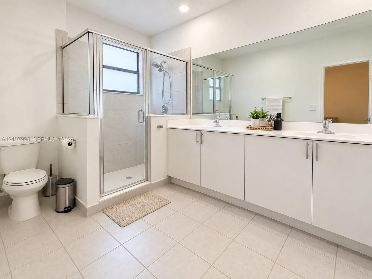 Dual-sink vanity with white cabinetry and solid surface countertop, large wall-mounted mirror, glass-enclosed shower with tile surround, and tile flooring