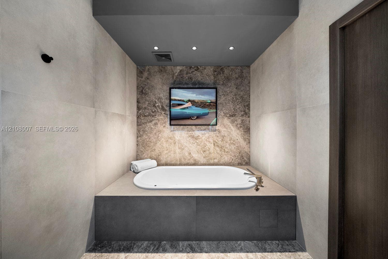 Built-in drop-in bathtub featuring a dark stone-finish base, large-format matte wall tiles, natural stone accent wall, and recessed ceiling lighting