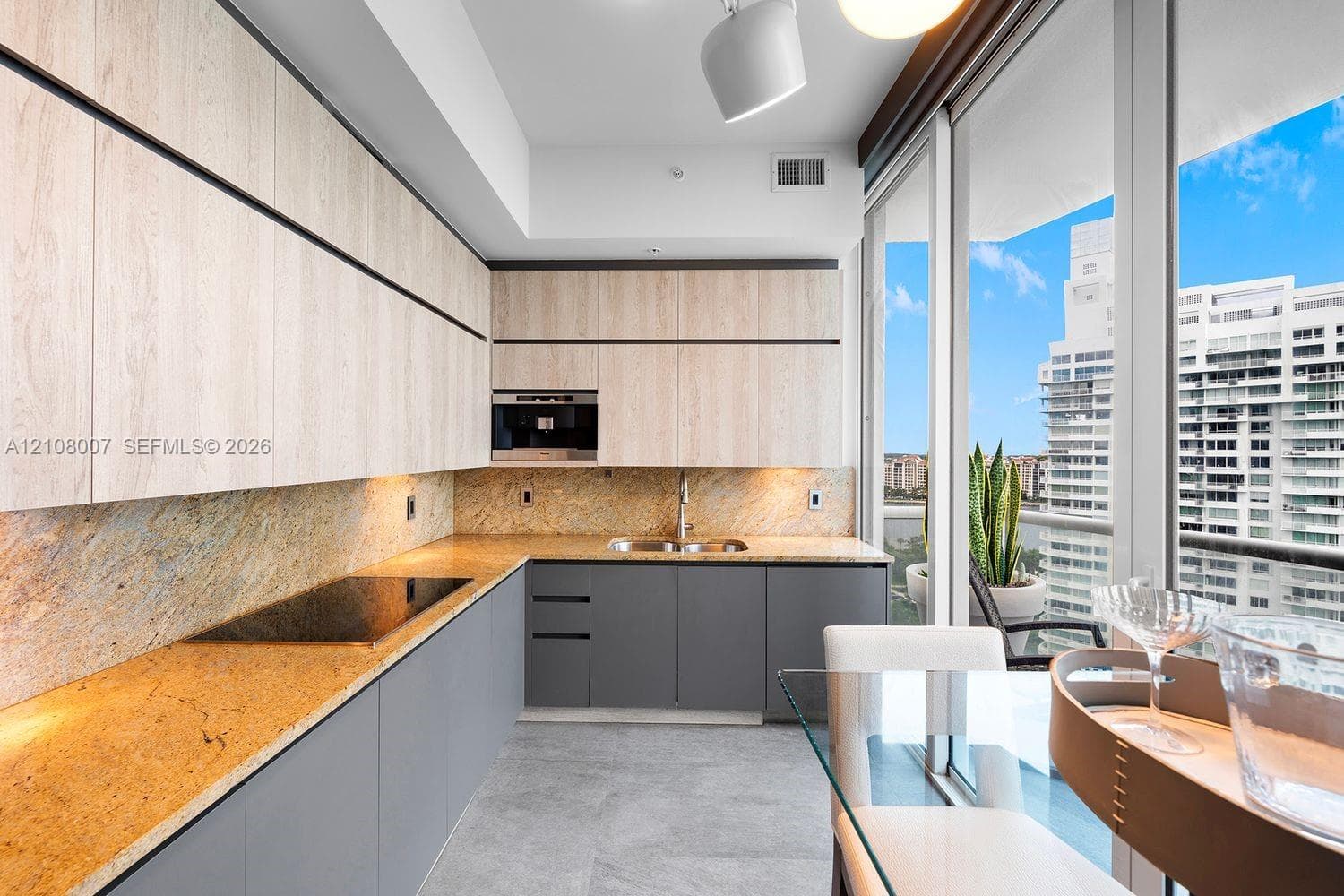 Kitchen featuring light wood-finish cabinetry, gray base cabinets, stone countertops with a matching backsplash, integrated cooktop, and floor-to-ceiling glass sliding doors