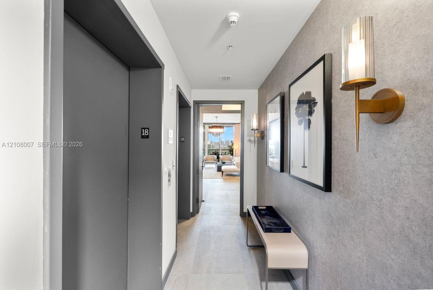 Hallway featuring textured grey wall cladding, gold-toned metallic wall sconces with cylindrical glass shades, and light-toned tile flooring