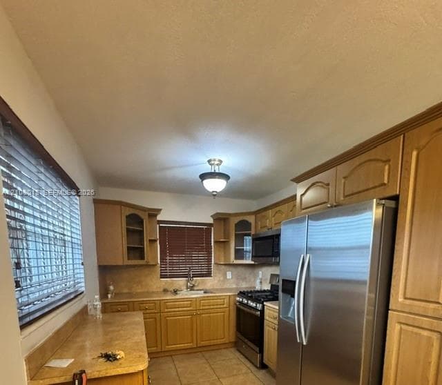 Kitchen featuring light-finish wood cabinetry, integrated stainless steel appliances, neutral stone-finish countertops, tiled flooring, and a flush-mount overhead light fixture