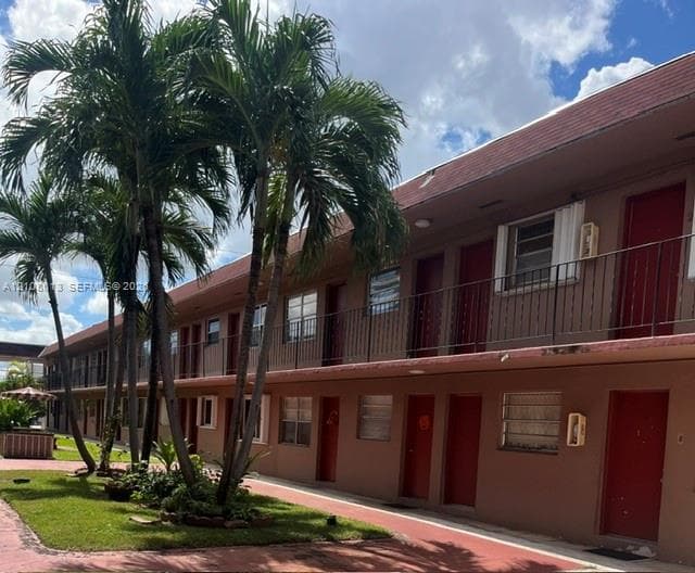 Multi-story exterior featuring a stucco facade, second-level metal balcony railings, prominent palm trees, and landscaped green space