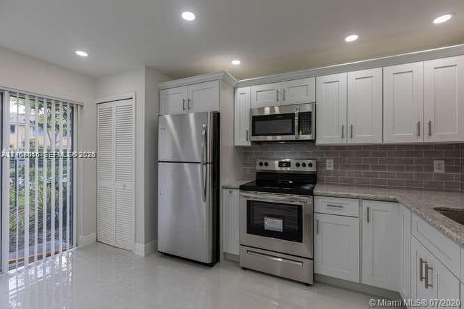 Kitchen featuring white shaker cabinetry, stainless steel appliances, neutral stone-finish countertops, subway tile backsplash, and polished tile flooring