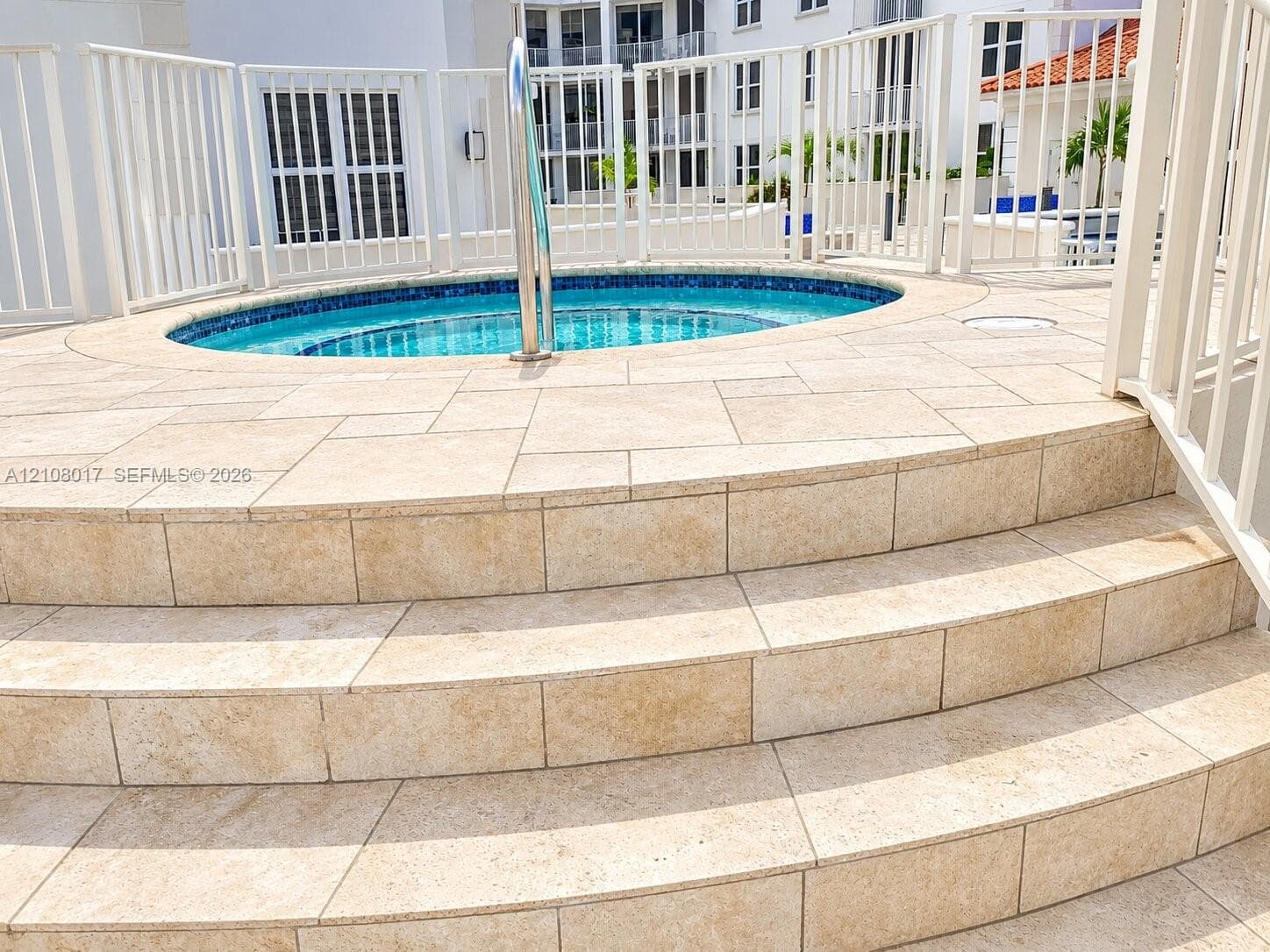Elevated spa featuring a blue tiled interior, metal handrail, surround of stone-finish paver decking, and white perimeter fencing