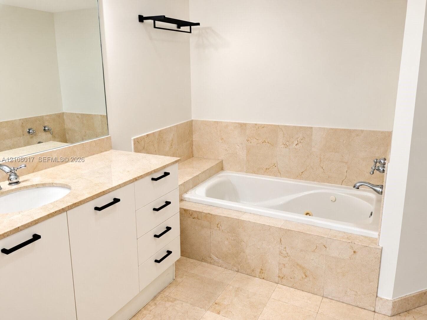 Bathroom featuring a drop-in jetted tub with stone tile surround, stone-topped vanity with undermount sink, white cabinetry with matte black hardware, and a wall-mounted towel rack
