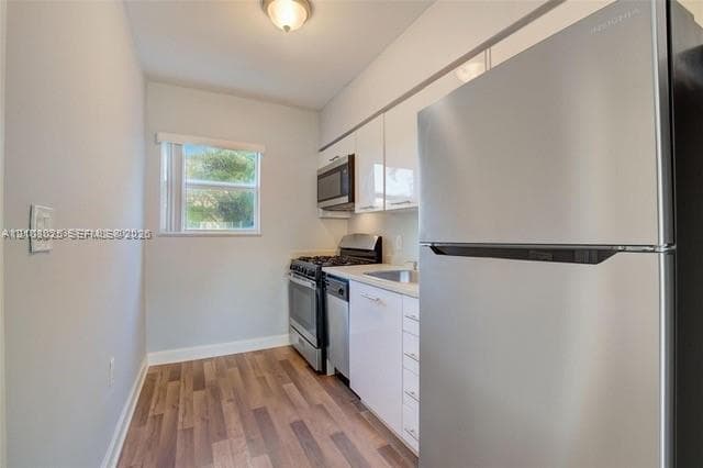 Kitchen featuring glossy white flat-panel cabinetry, stainless steel appliances, wood-finish flooring, and a window providing natural light