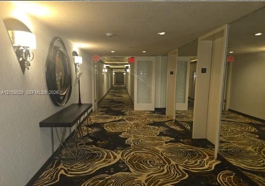 Hallway featuring patterned wall-to-wall carpeting, decorative wall-mounted sconces, textured wall coverings, and mirrored paneling