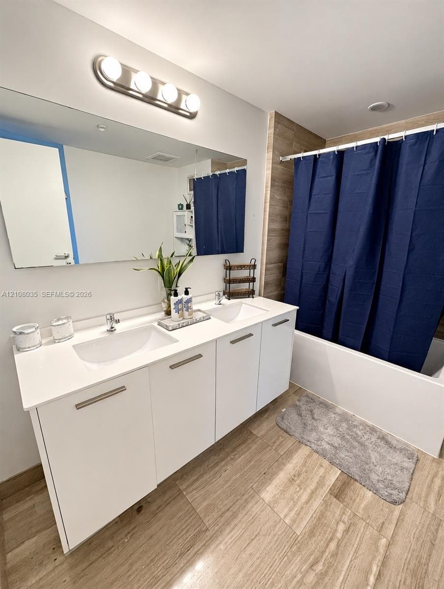 Dual-sink vanity with a solid-surface countertop, full-width vanity mirror, four-light wall fixture, and tub-shower combination with floor-to-ceiling tile surround