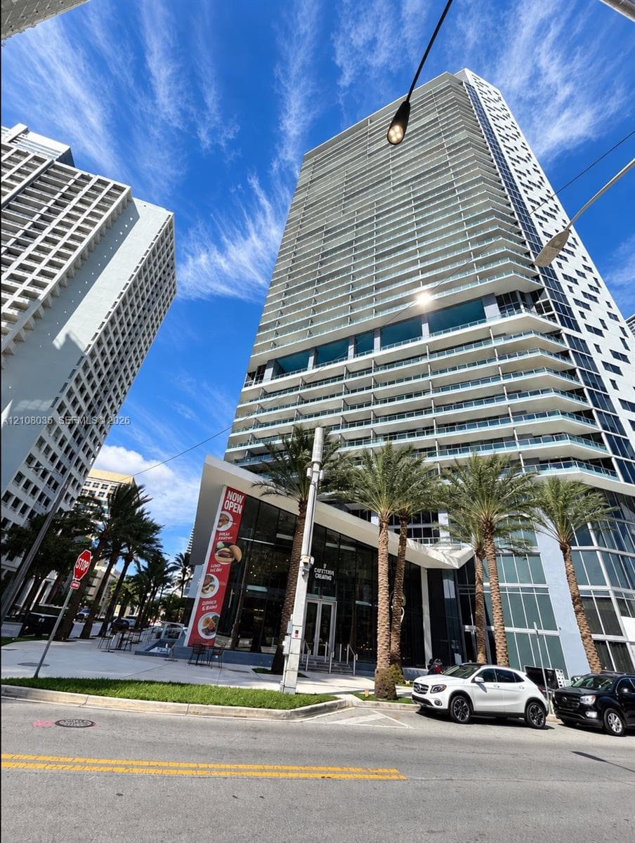 High-rise residential tower featuring glass-panel balconies, floor-to-ceiling windows, and ground-level storefronts with modern angular architectural accents