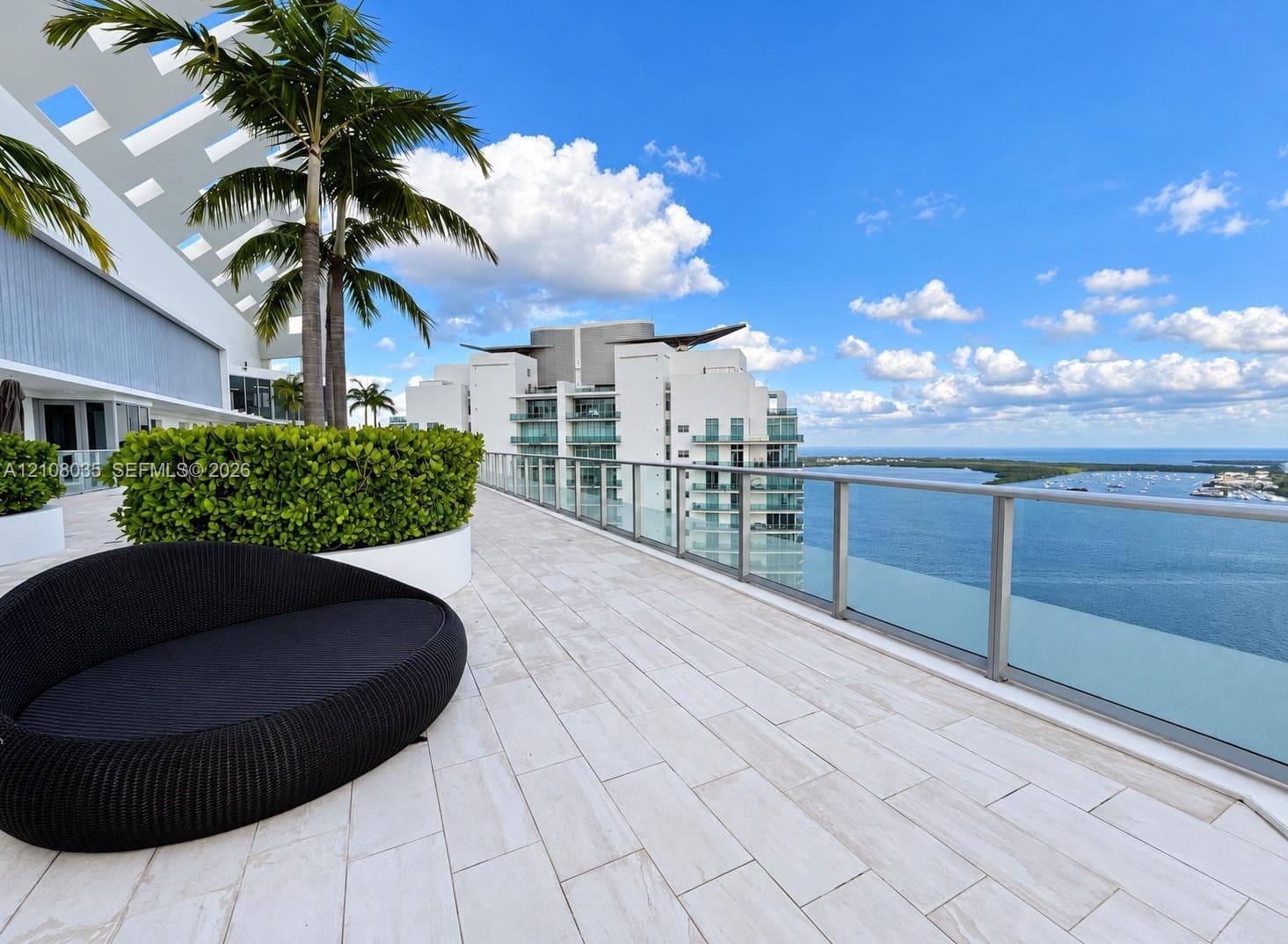 Expansive terrace featuring light-toned tile flooring, built-in planters with manicured greenery, and a glass-paneled railing overlooking the water