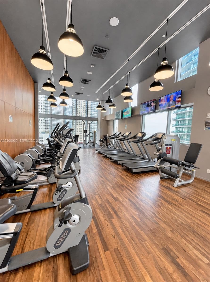 Fitness center featuring wood-finish flooring, floor-to-ceiling windows, panelled wood accent wall, and industrial-style pendant lighting