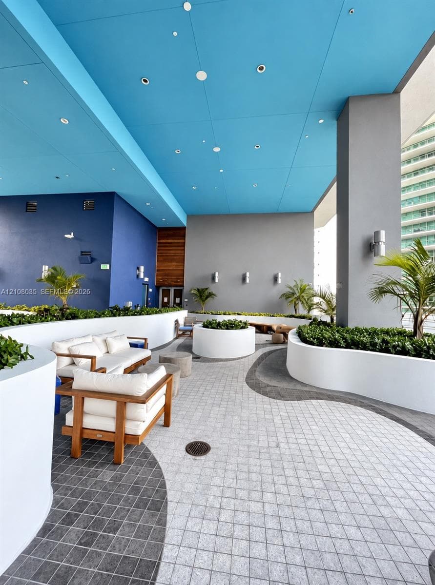 Outdoor covered terrace featuring curvilinear white concrete planters, two-tone patterned stone tile flooring, recessed ceiling lighting, and wall-mounted exterior sconces
