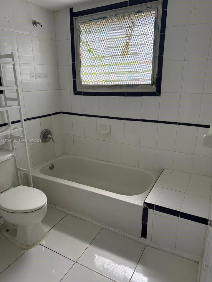 Bathroom featuring white tile flooring and wall tiling with dark accent trim, built-in tiled tub surround, integrated soaking tub, and a window with a security screen