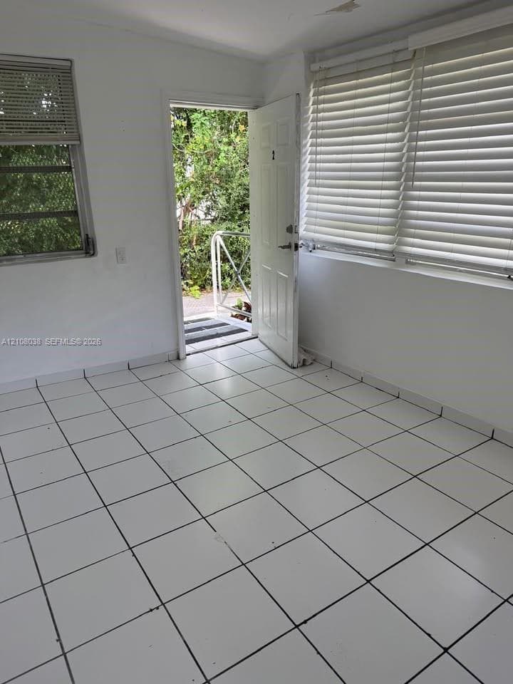 Interior space featuring white tile flooring, horizontal window blinds, and an exterior door opening to a paved pathway