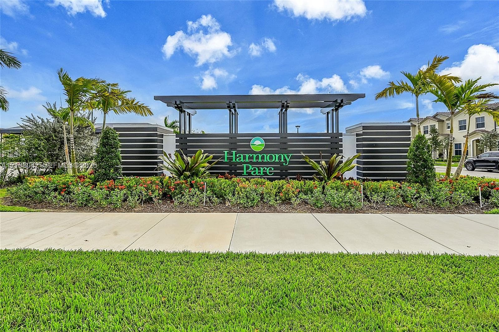 Community entrance monument featuring horizontal slat architectural detailing, integrated signage, concrete walkway, and professionally landscaped tropical foliage