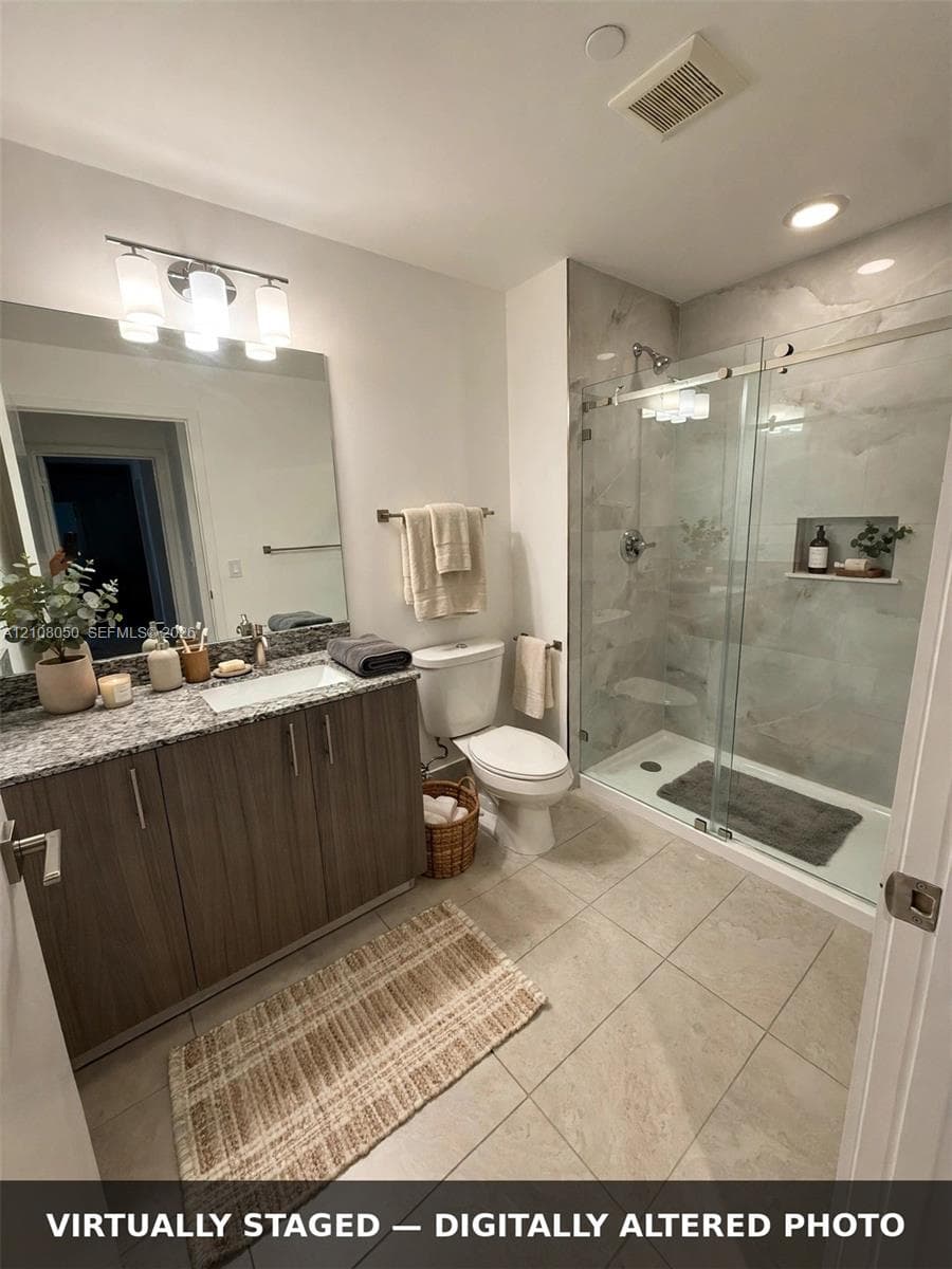 Bathroom featuring wood-finish cabinetry, stone-patterned countertop with an integrated rectangular sink, large format tile flooring, and a walk-in shower with glass sliding doors and a built-in recessed shelf
