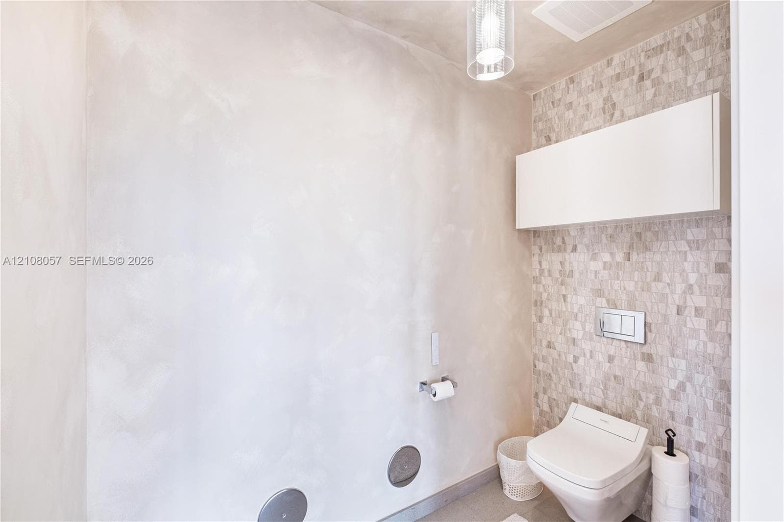 Wall-mounted toilet with a dual-flush actuator plate, mosaic tile accent wall, wall-mounted storage cabinet, and glass pendant lighting