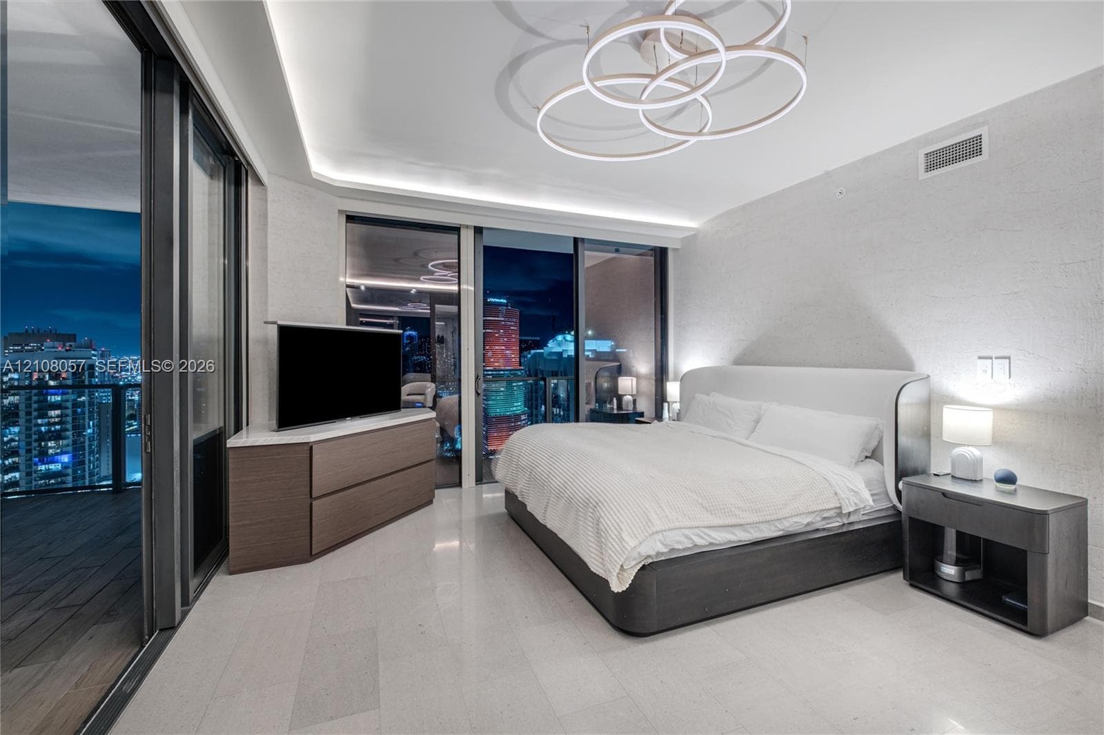 Floor-to-ceiling glass sliding doors, recessed cove lighting, sculptural ring chandelier, stone-finish tile flooring, and private balcony access