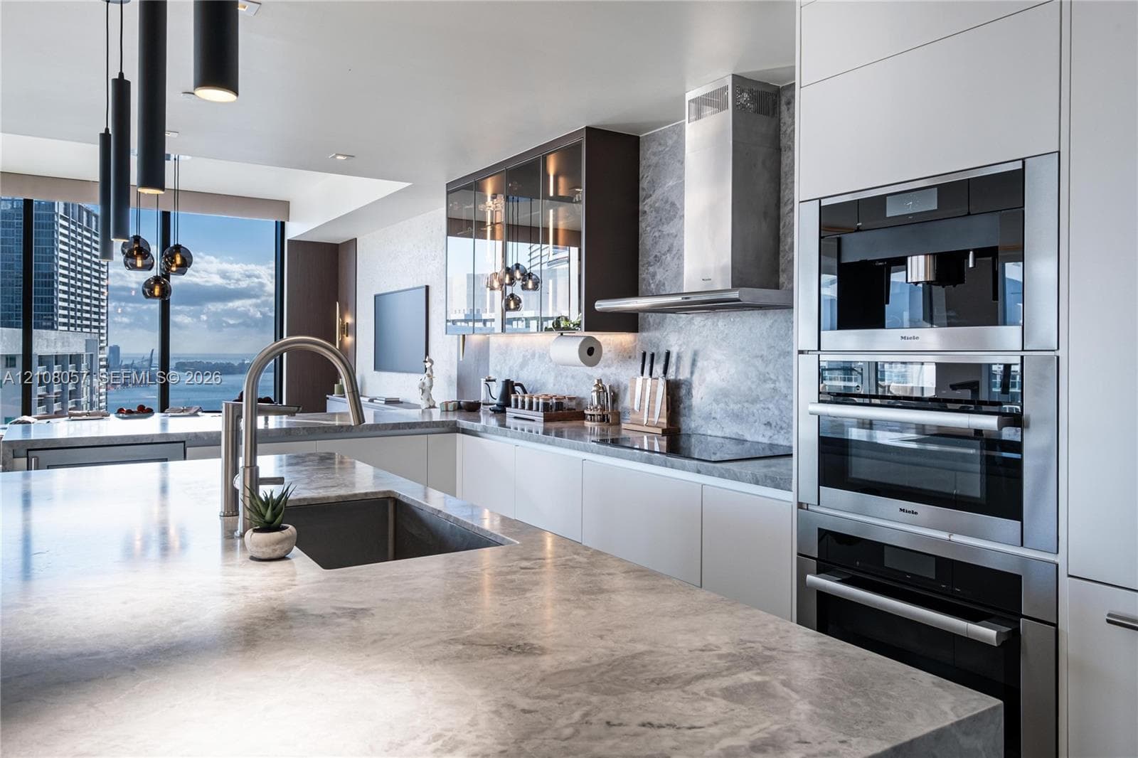 Contemporary kitchen featuring stone countertops, integrated stainless steel appliances, matte-finish flat-panel cabinetry, glass-front upper storage, and sleek pendant lighting