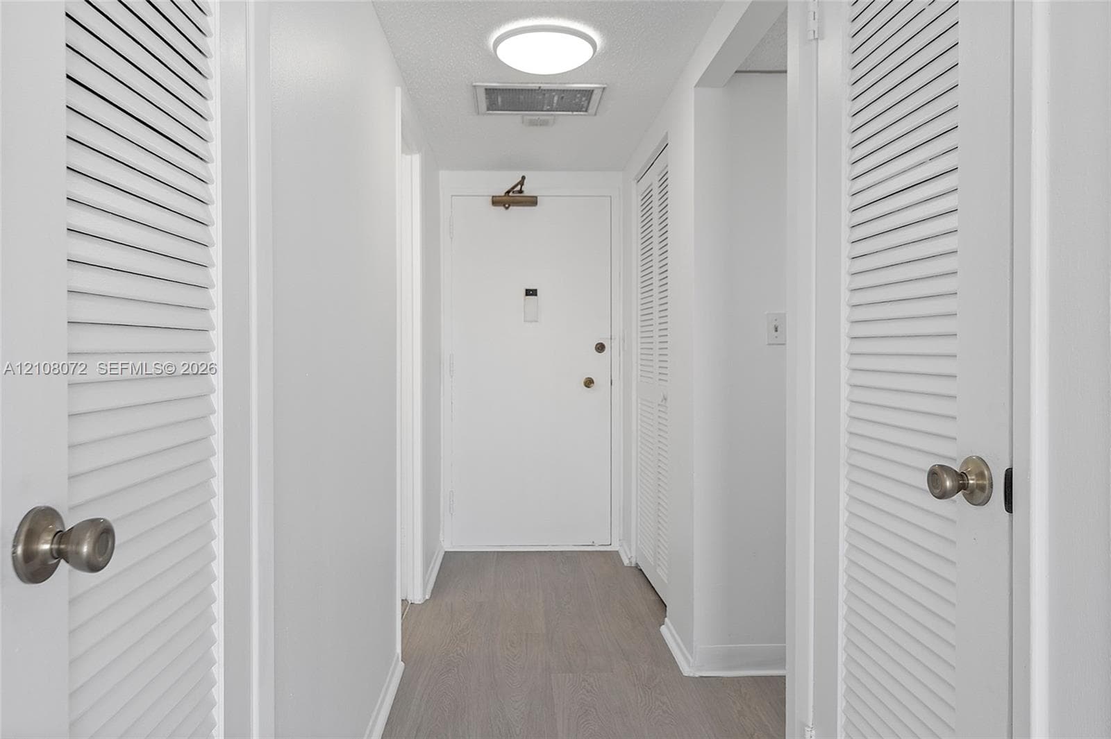 Interior hallway featuring wood-finish flooring, dual louvered closet doors with metallic hardware, and flush-mount ceiling lighting
