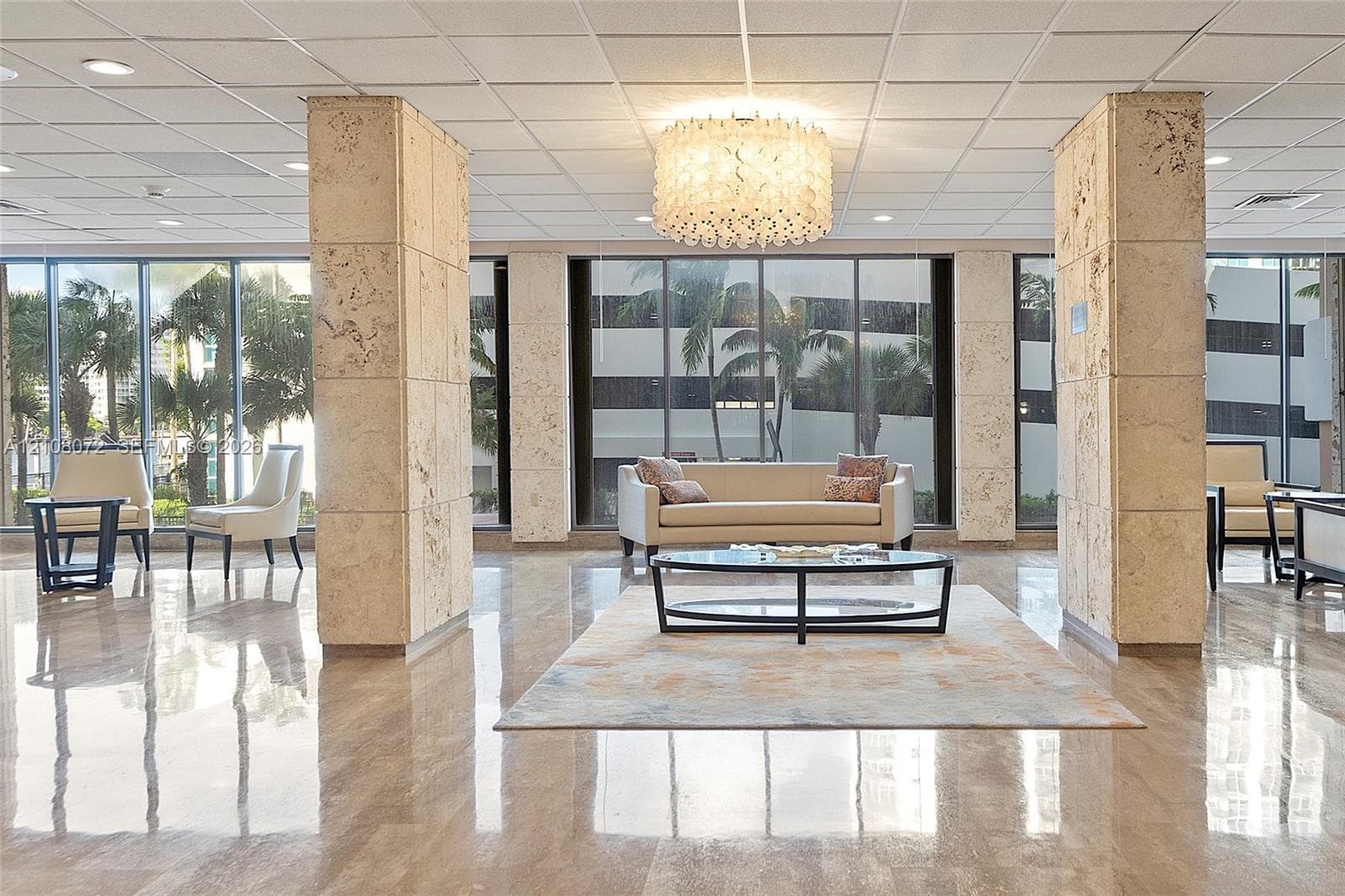 Spacious lobby featuring polished stone flooring, floor-to-ceiling windows, architectural stone-clad support pillars, a multi-tiered circular chandelier, and a drop ceiling