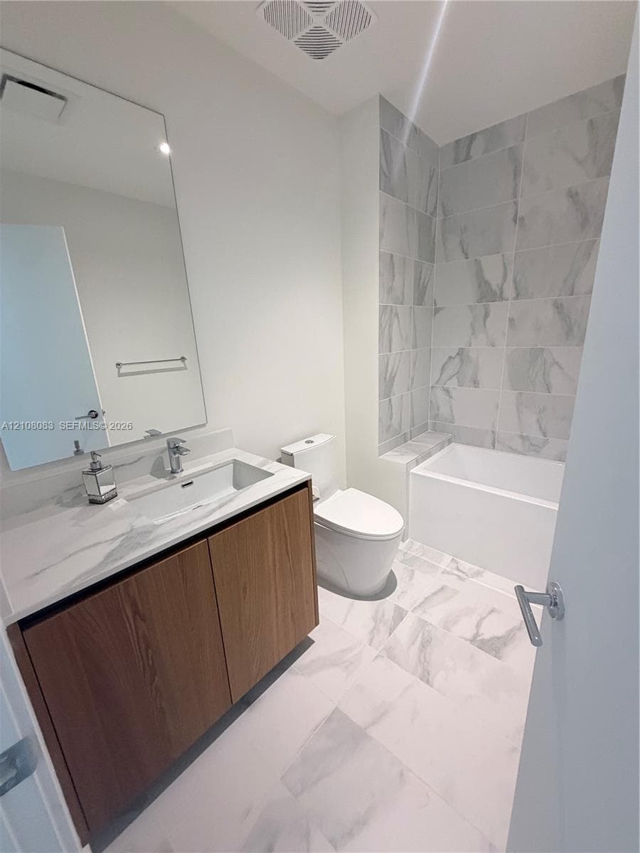 Bathroom featuring a wood-finish floating vanity, stone-finish countertop with integrated sink, large-format tile flooring, and a bathtub enclosure with floor-to-ceiling stone-finish tile surround